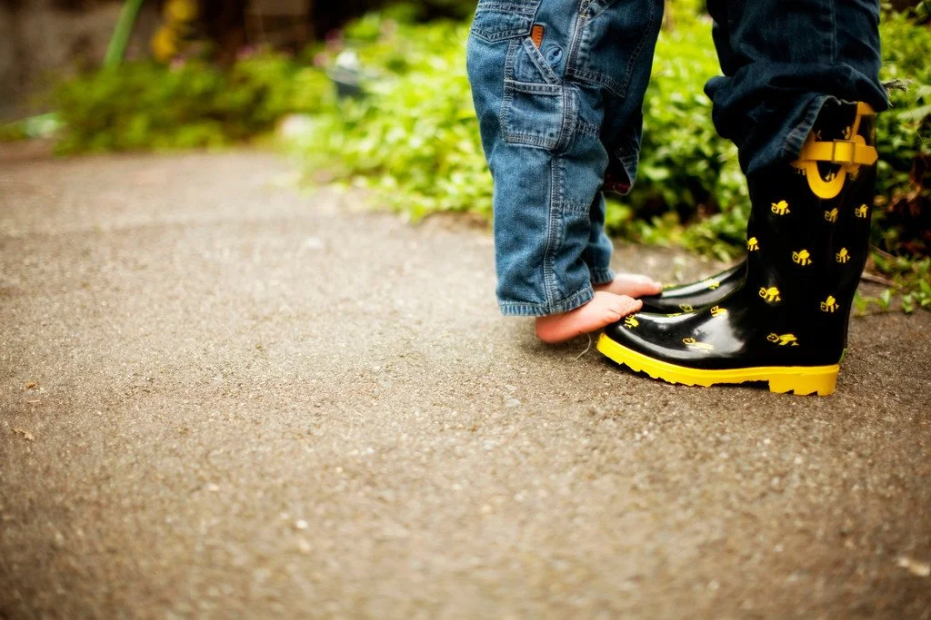 Child wearing jeans and an oversized black rain boot with yellow airplane pattern helps a toddler in a yellow rain boot with black soles stand on the sidewalk.