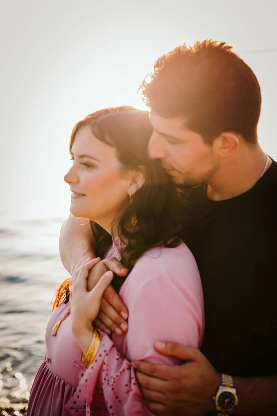 A man and woman embrace near the beach at sunset, with the sun setting behind them.