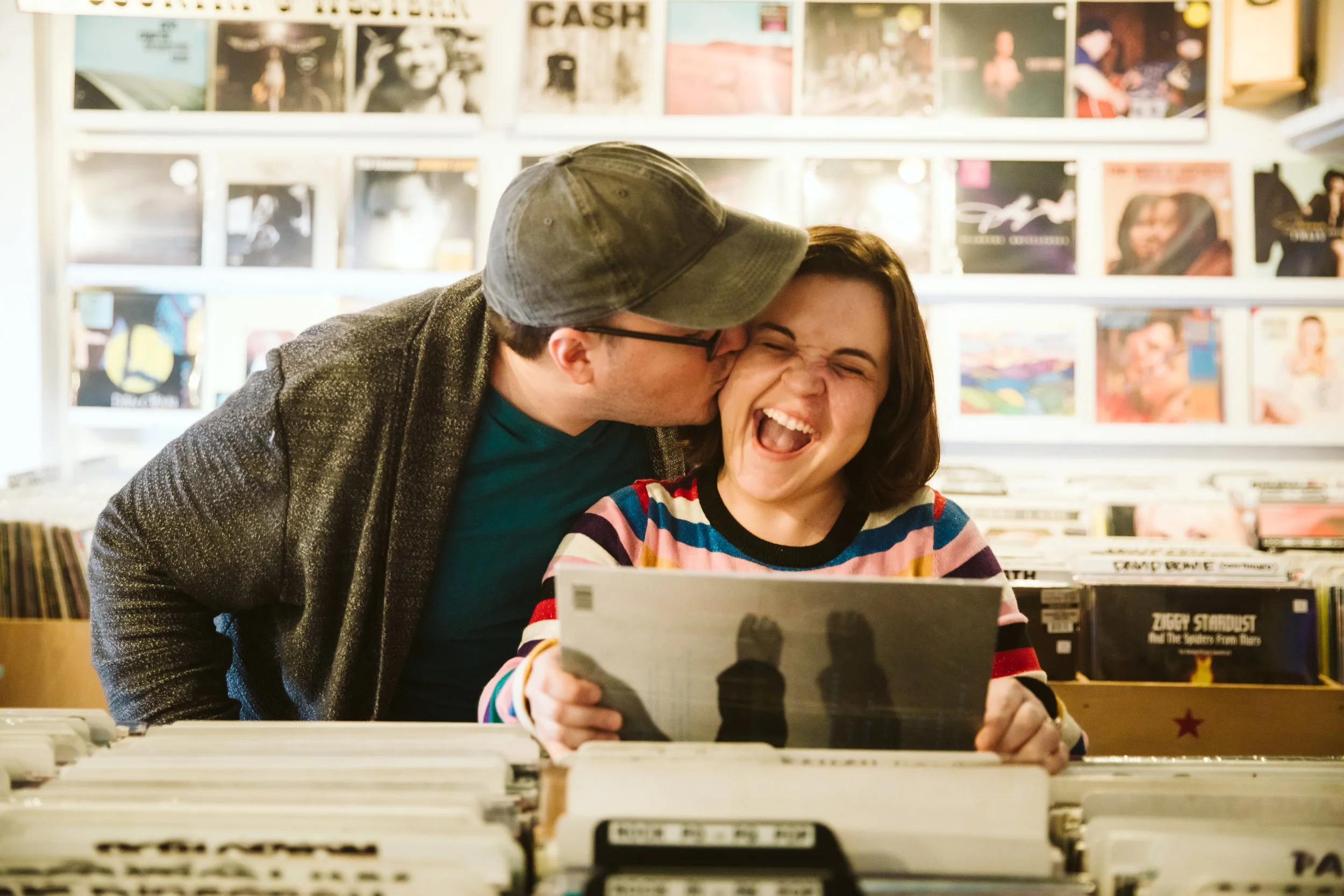 A man is kissing a woman on the cheek while she laughs and looks at a record album inside a record store, with vinyl records and album covers visible in the background.