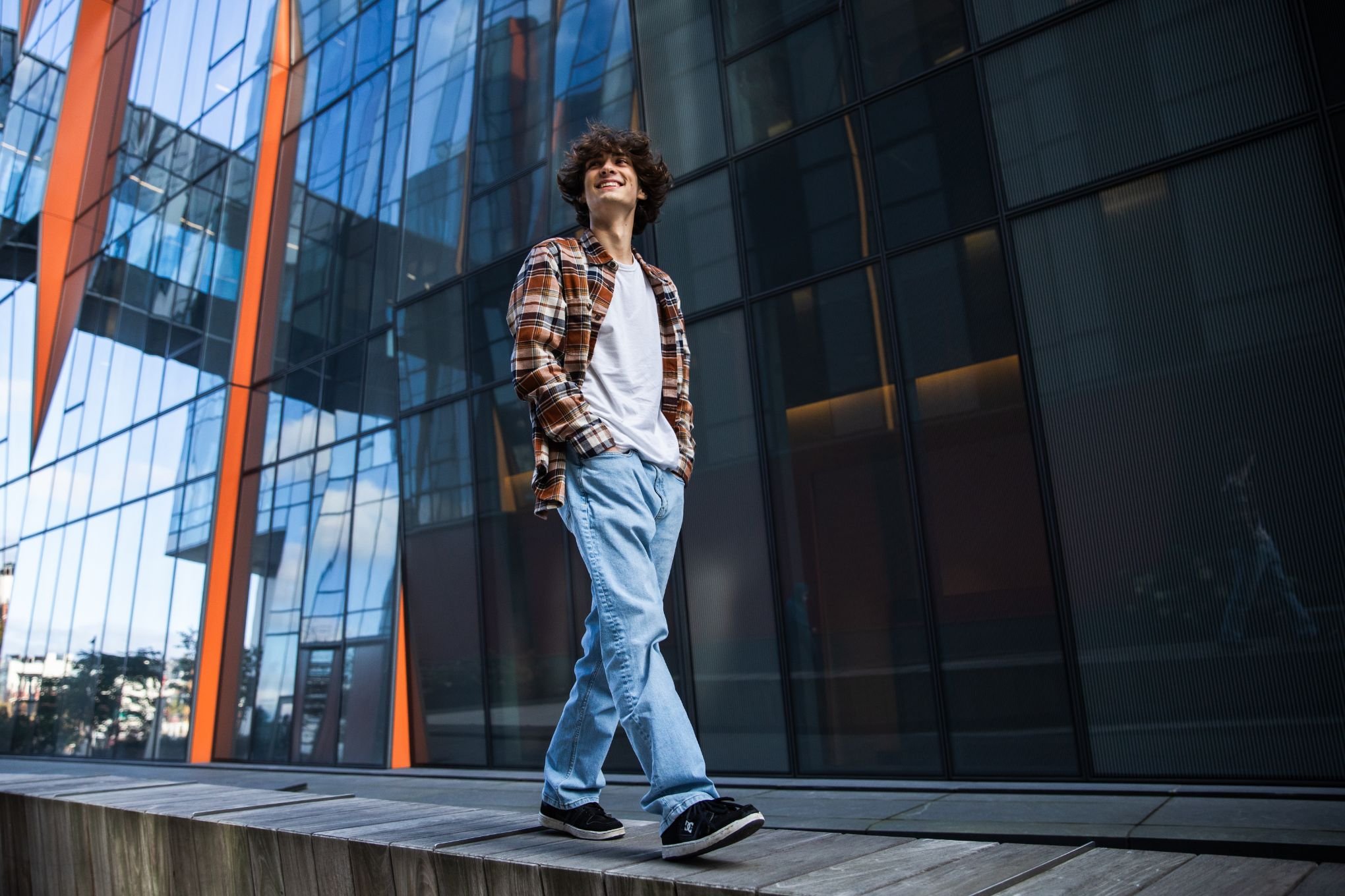 Young man walking outdoors near a modern glass building, smiling and wearing casual clothes including a plaid shirt, white t-shirt, and jeans.
