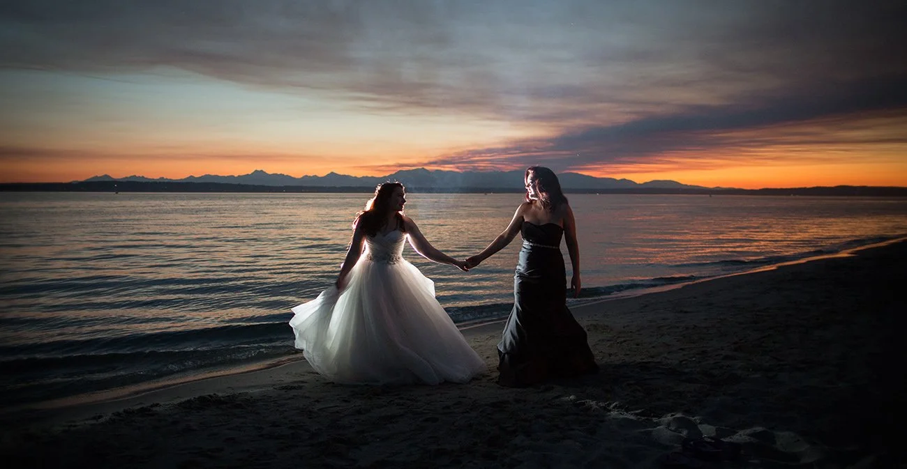 Two women holding hands on the beach during sunset, one in a white wedding gown and the other in a black dress.