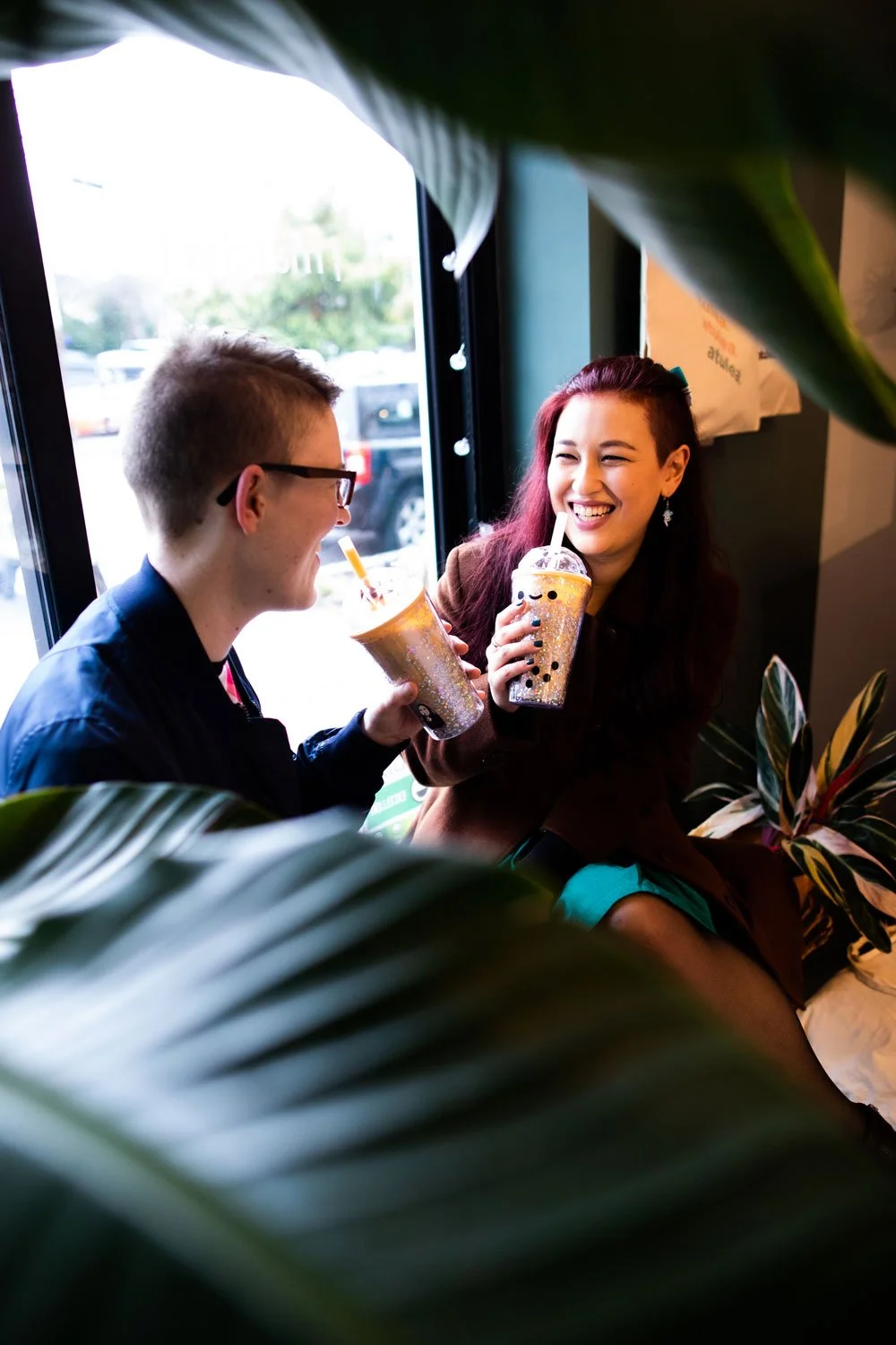 Two friends, a man with glasses and a woman with long dark hair, are smiling and enjoying drinks inside a cafe near a window, framed by large green plants.