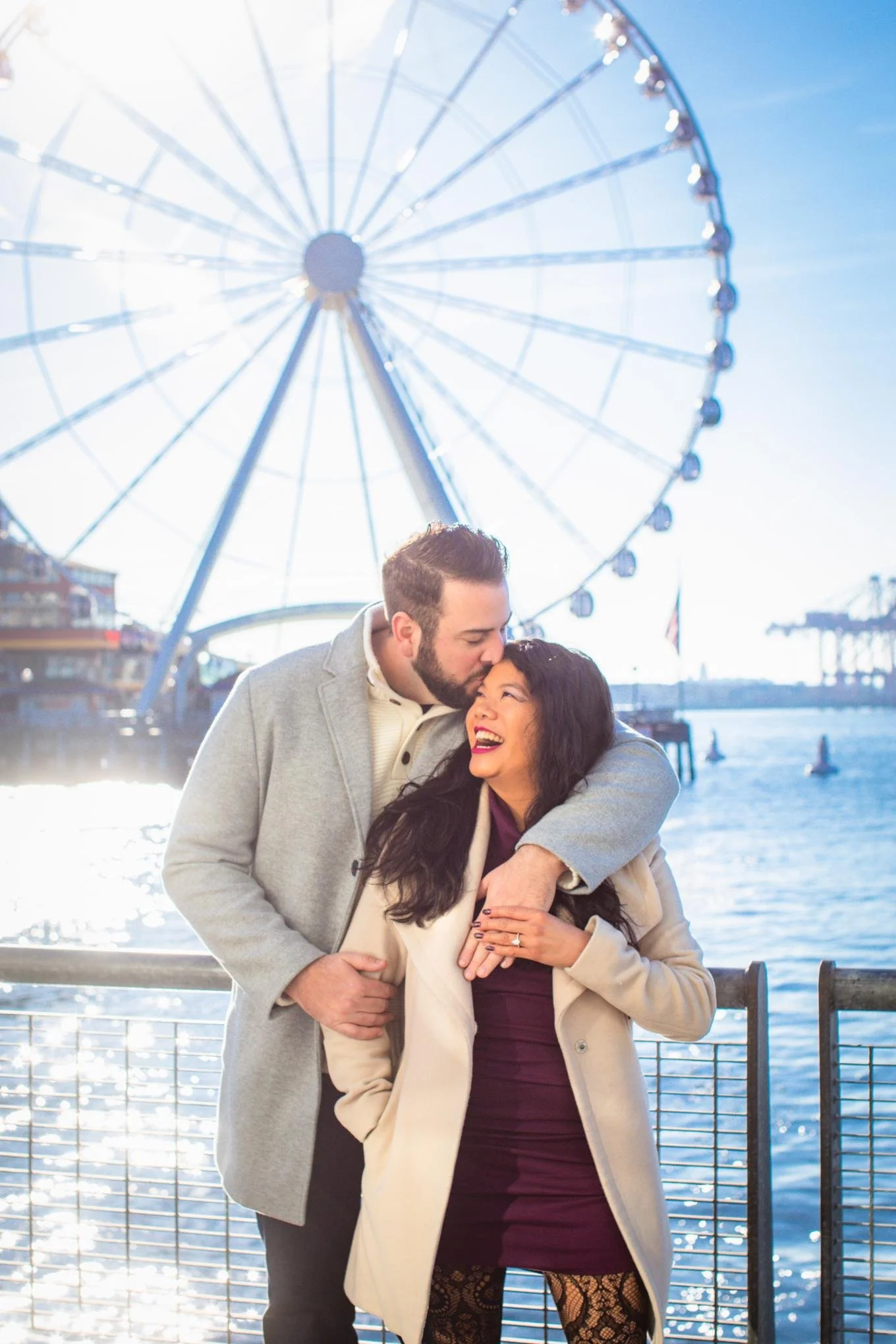 A couple sharing a romantic moment near a waterfront with a large Ferris wheel in the background.