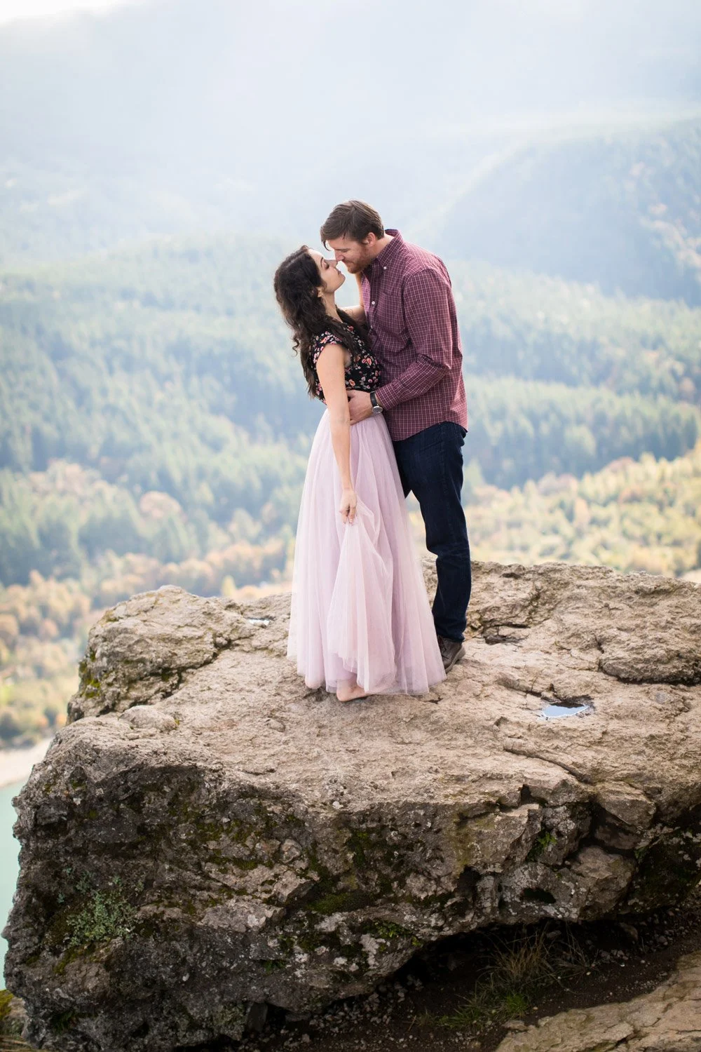 A couple kissing on a rock overlooking mountains and a valley, with a rainbow in the background.