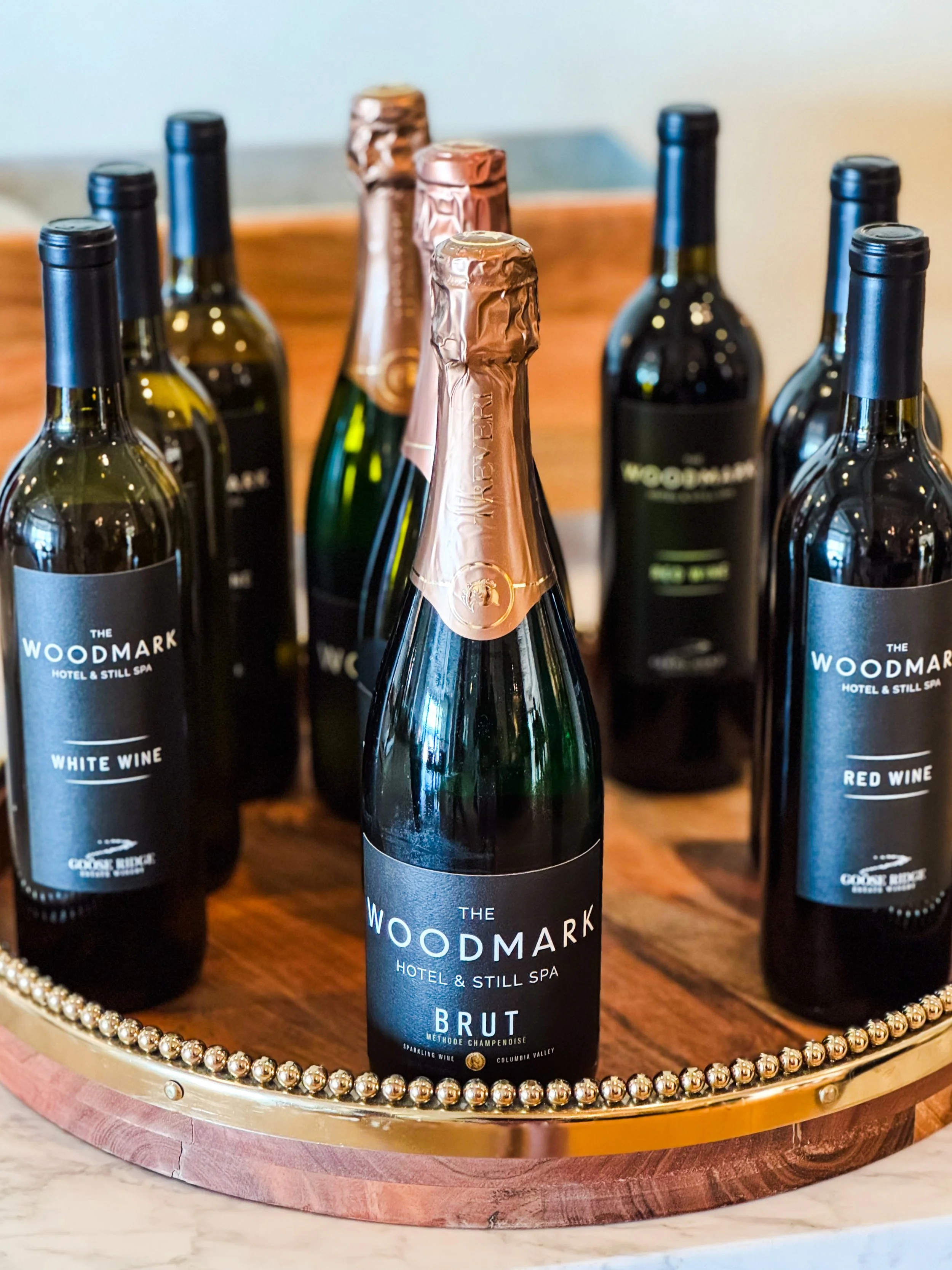 A tray of bottles of wine, including a bottle of The Woodmark Hotel & Still Spa Brut with a pink foil top, surrounded by bottles of red and white wine, on a wooden tray with a gold beaded edge.
