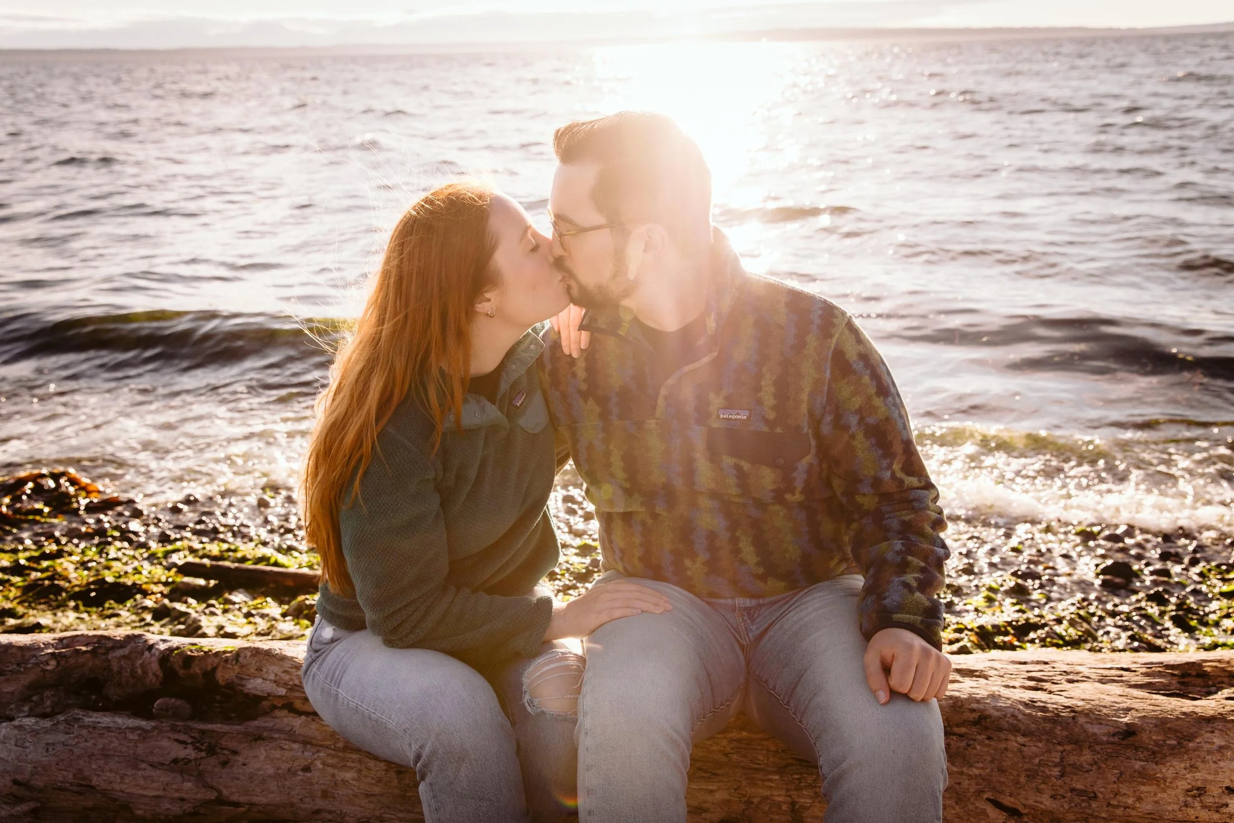 A couple sitting on a log by the water, sharing a kiss against the sunset over the ocean.