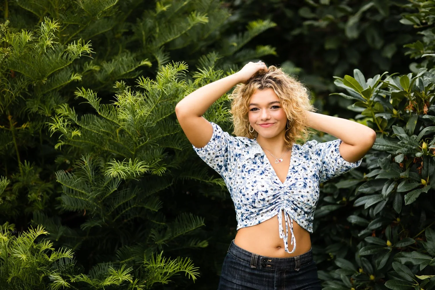A young woman with curly blonde hair smiling outdoors surrounded by green plants, wearing a floral crop top and dark jeans.