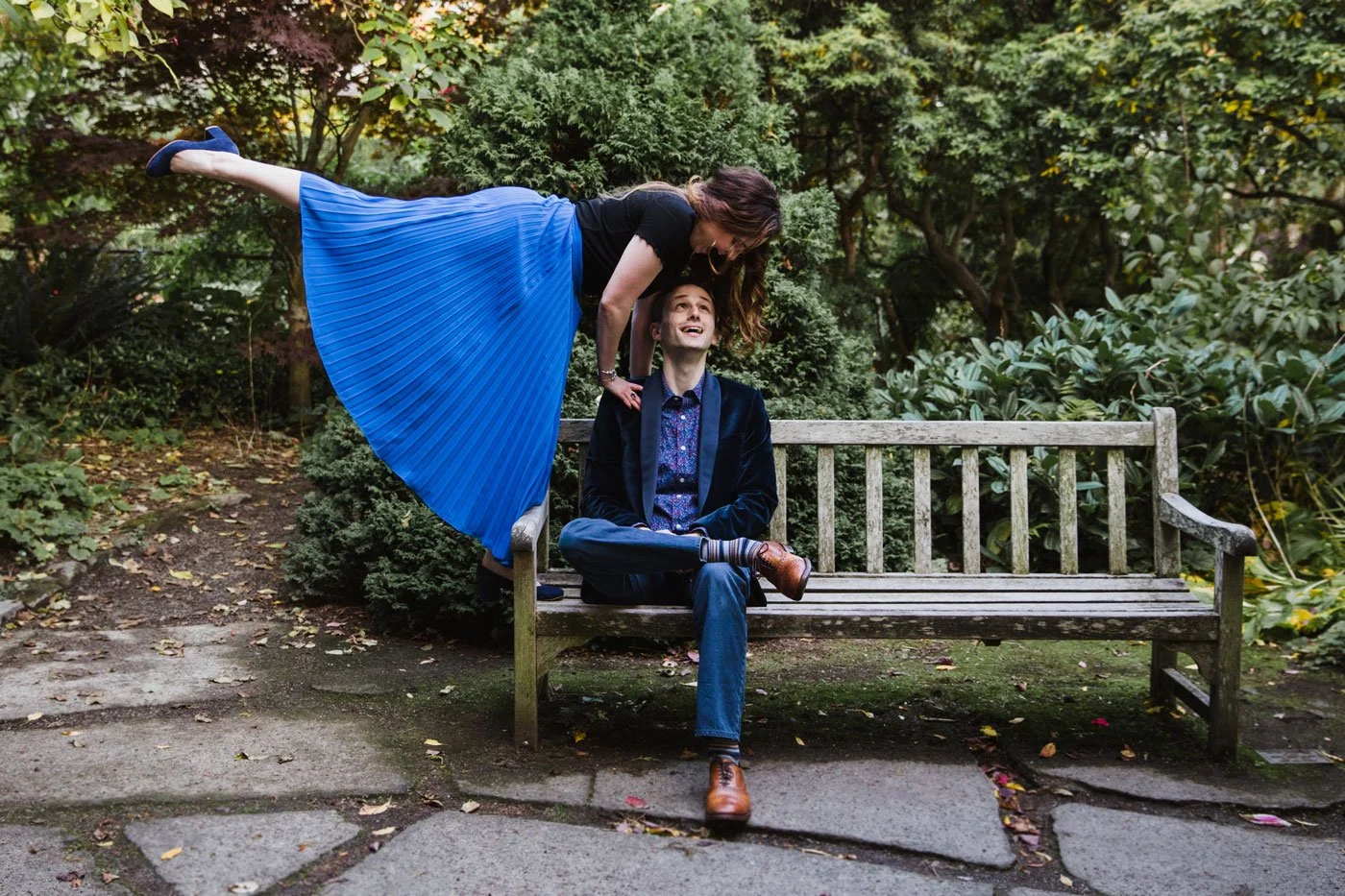 A woman in a blue pleated skirt and black top is leaning over a seated man in a dark blazer and jeans, with her arm around his shoulder, both smiling in a garden setting.