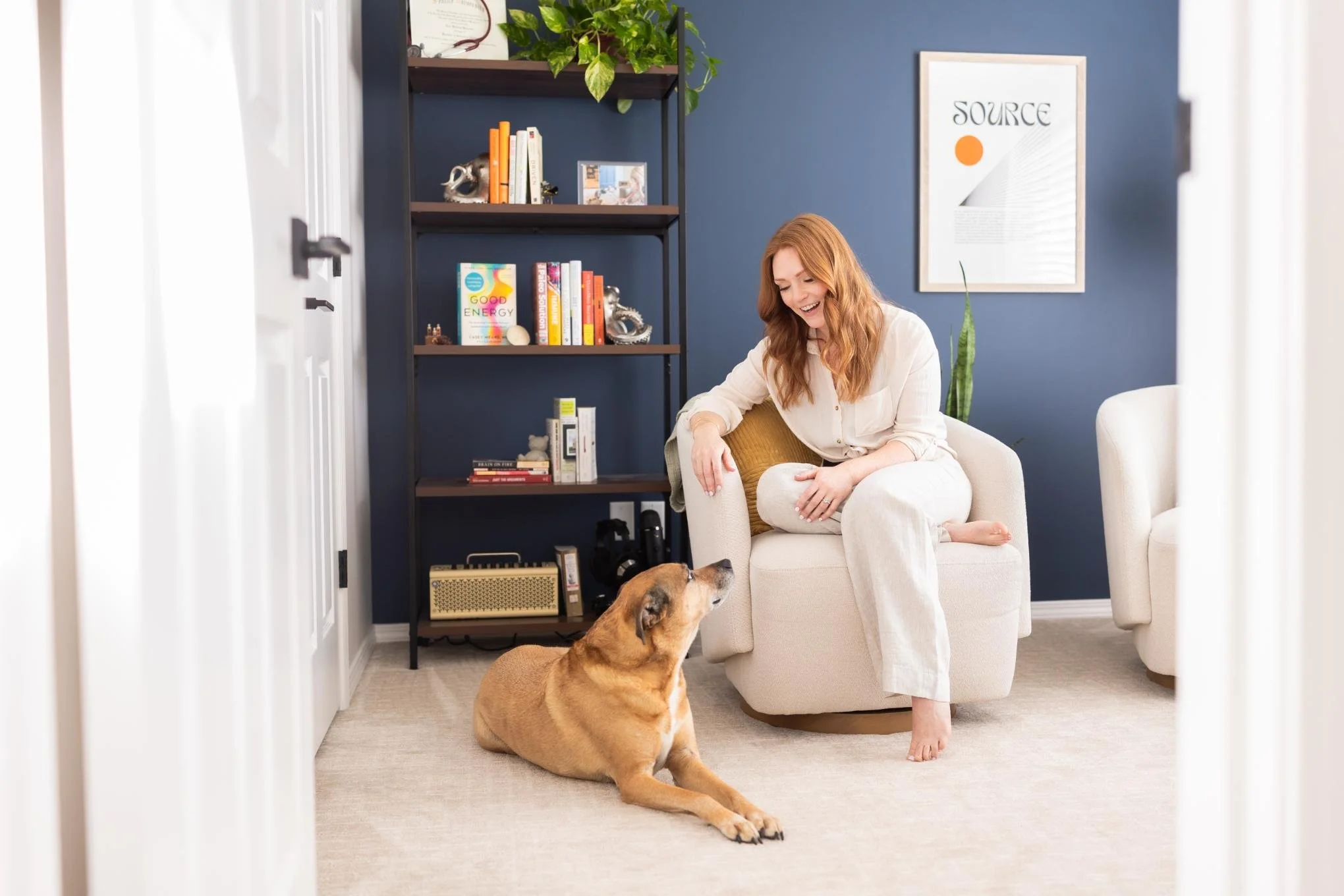 A branding photo of woman with red hair and light-colored pajamas sitting on an armchair, playing with a brown dog lying on the carpet in a living room with a blue accent wall, a framed quote, a bookshelf with books and decorative items, and plants.