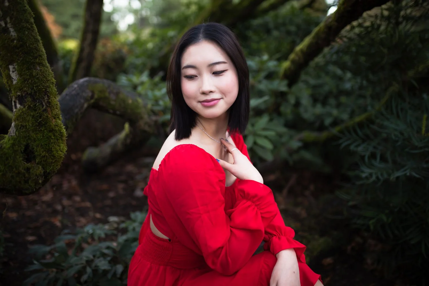 A woman with short black hair wearing a red long-sleeve dress sitting outdoors in a lush green forest, with her eyes closed and a gentle smile, touching her chest with her left hand.