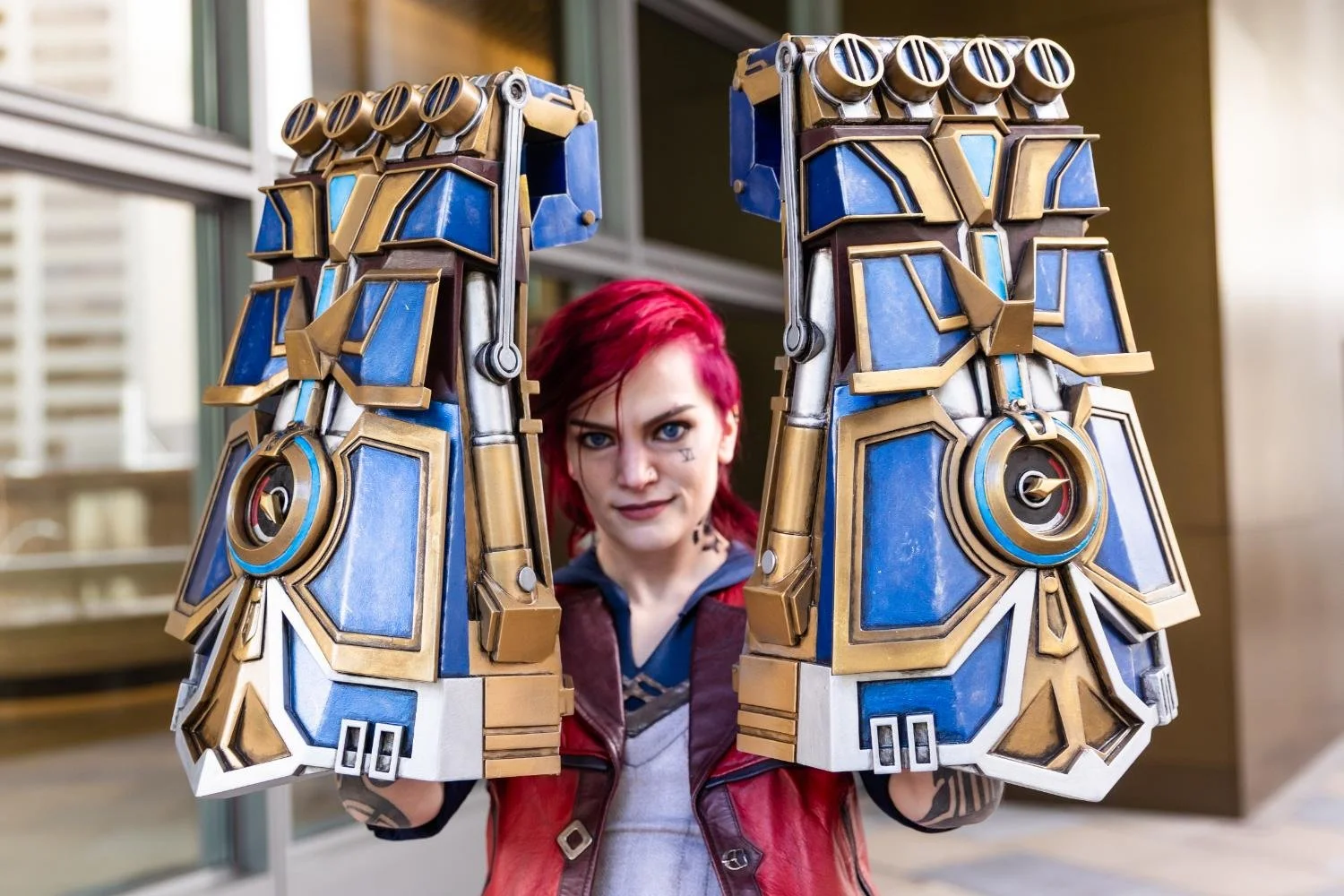 Person with red hair in cosplay costume holding large, detailed, sci-fi styled blue and gold robotic gauntlets.