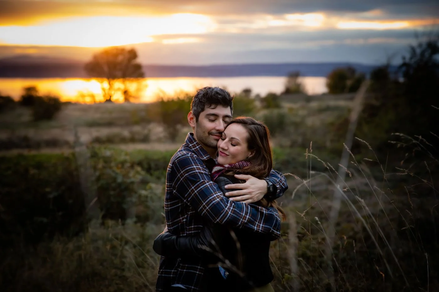 A young couple embracing outdoors during sunset, with a scenic landscape of trees and water in the background.