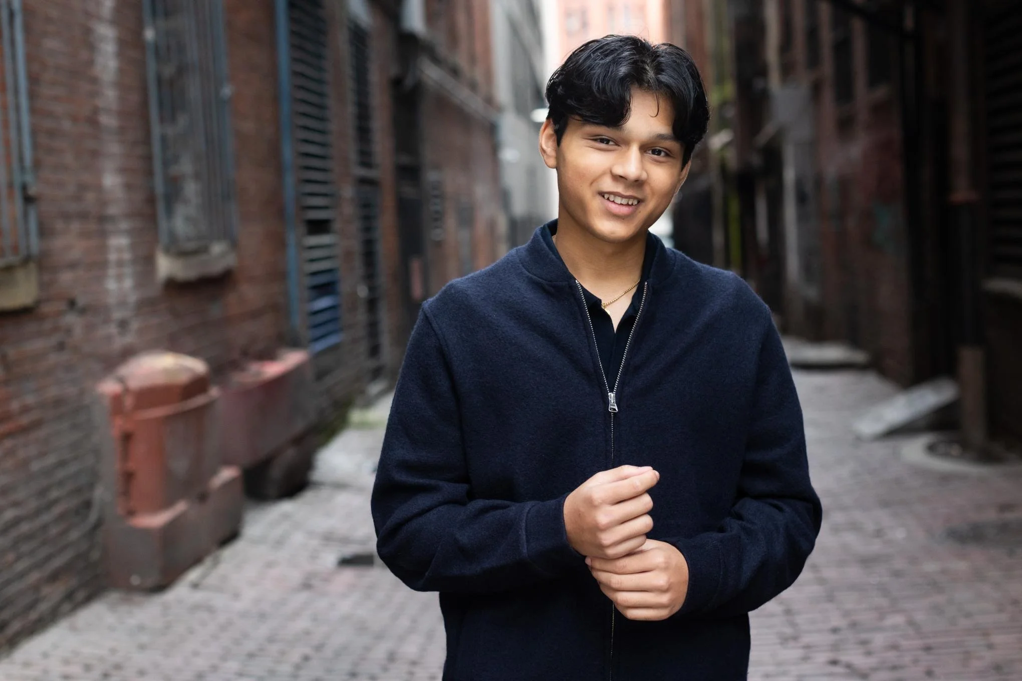 A young man with black hair in a navy blue jacket smiling and standing in an alleyway with brick walls and windows.