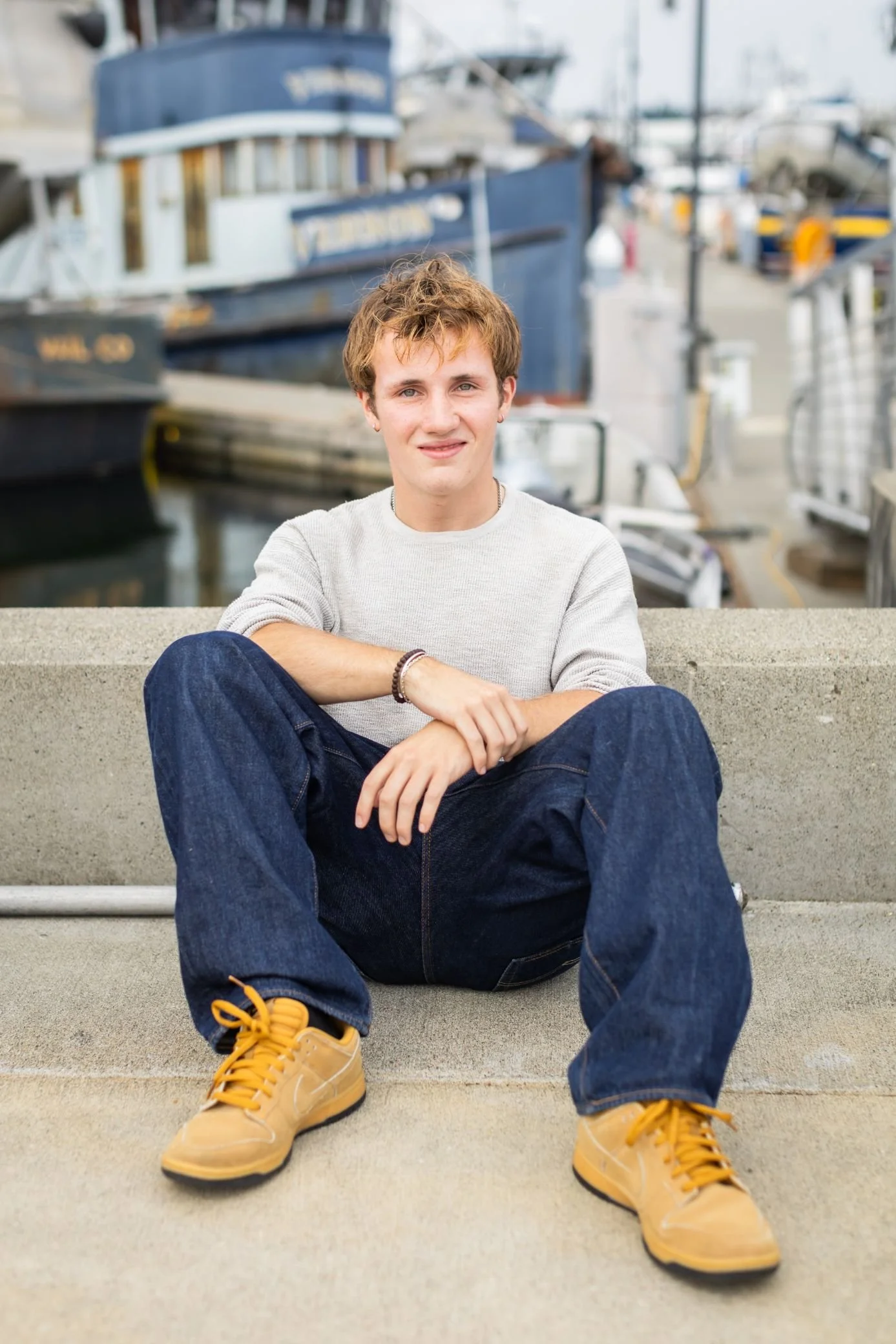 A young man sitting on a concrete ledge near a marina with boats in the background.