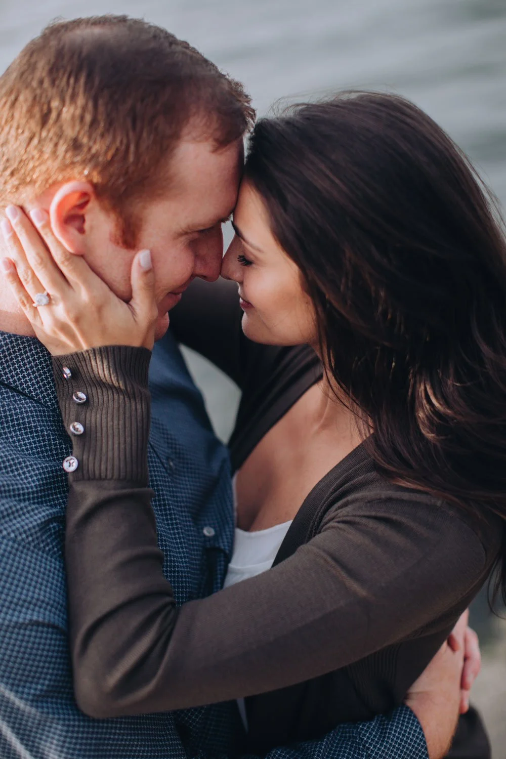 A romantic couple embraces closely with their foreheads touching, smiling, outdoors by the water.