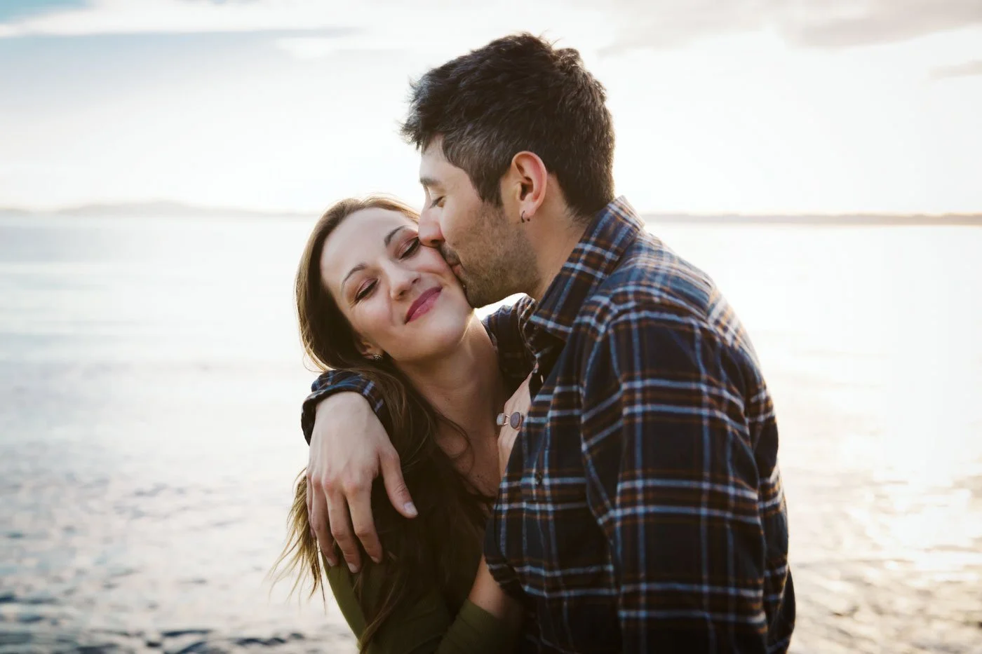 A couple embracing at the beach with the man kissing the woman's cheek and the woman smiling with her eyes closed.
