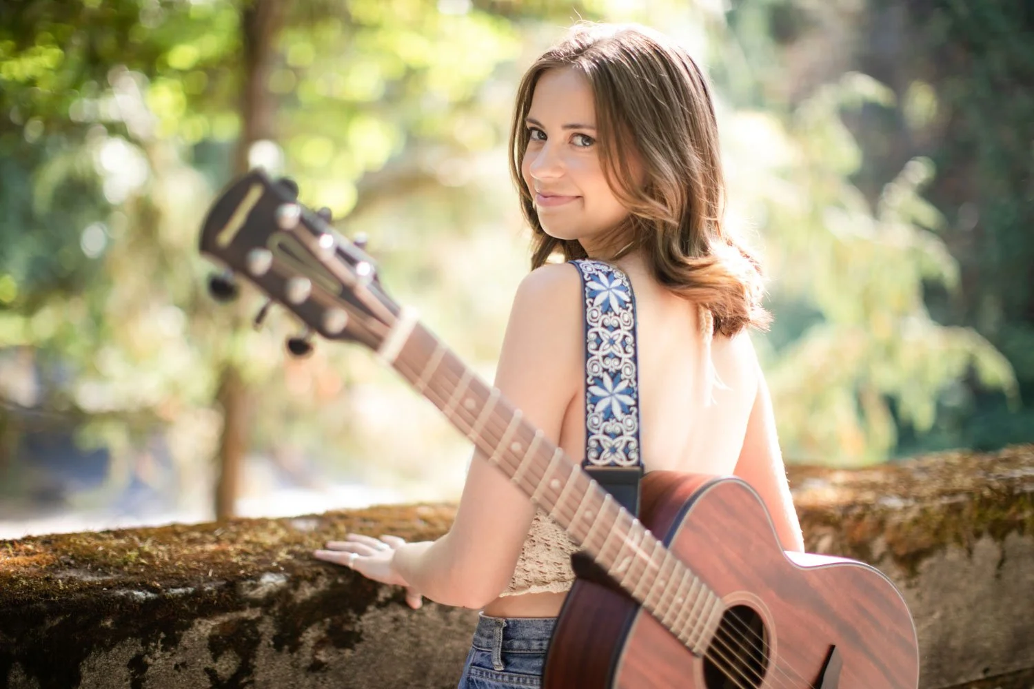 A young woman with brown hair holding an acoustic guitar while standing outdoors in a forested area, smiling at the camera.