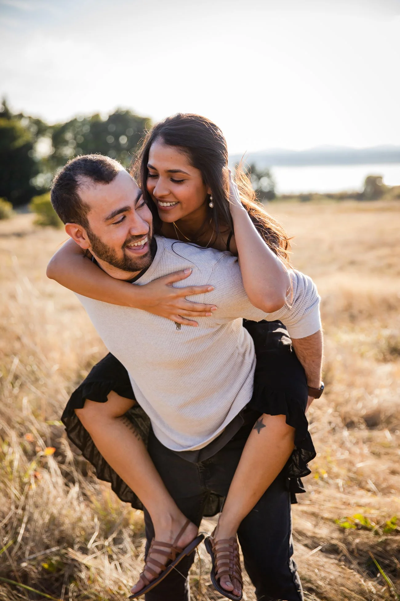 A young couple enjoying a playful moment outdoors during golden hour, with the woman on the man's back, smiling and embracing in a grassy field.