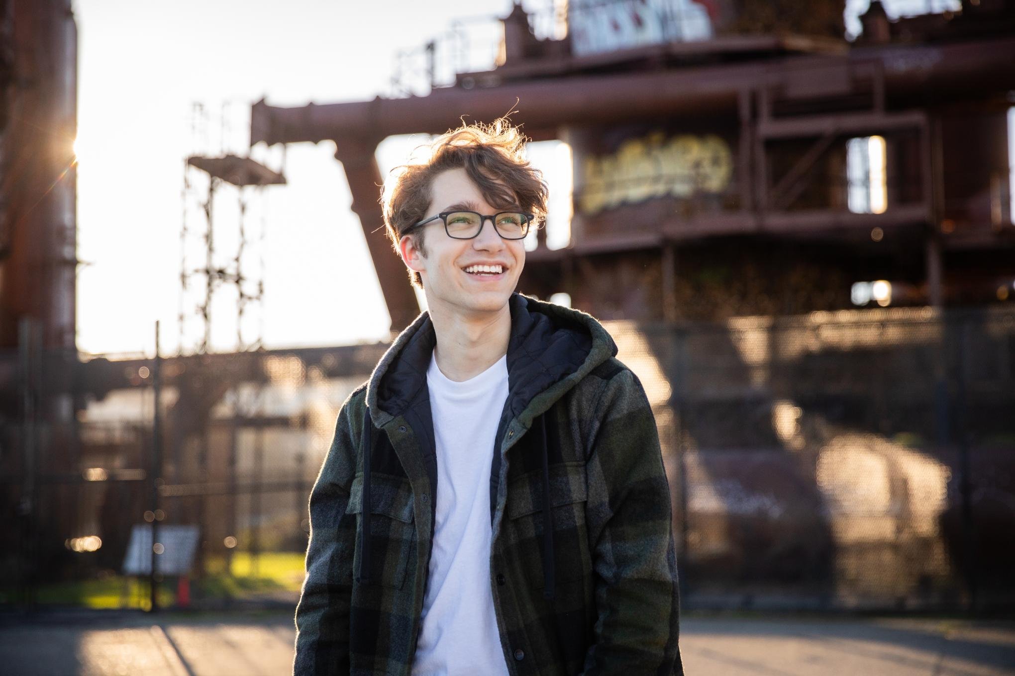 A young man with glasses and curly hair wearing a hoodie and plaid jacket outdoors during sunset, with an industrial structure in the background.