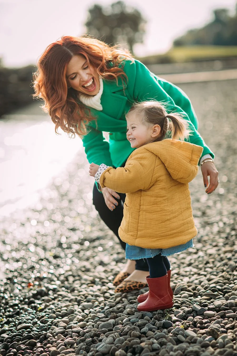 A woman and a young girl are outdoors on a pebble beach, laughing and playing together. The woman has red hair and is wearing a bright green coat, while the girl wears a yellow jacket and red rain boots.