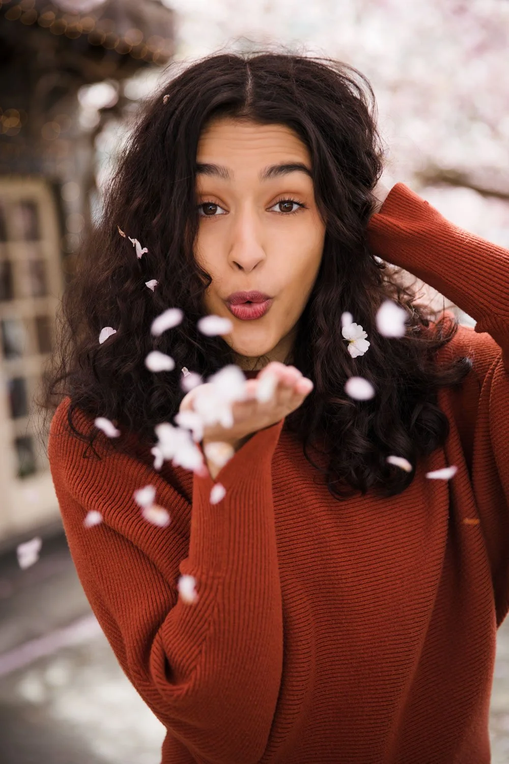 A young woman with curly dark hair wearing a rust-colored sweater is blowing flower petals towards the camera with a playful expression, outdoors with blurred background of trees and pink blossoms.