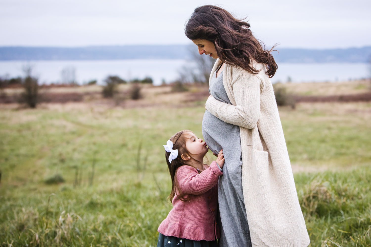 A pregnant woman in a beige cardigan is standing in a grassy field with her young daughter, who is wearing a pink sweater and dark skirt, hugging her belly and giving her a kiss, with a lake and trees in the background.