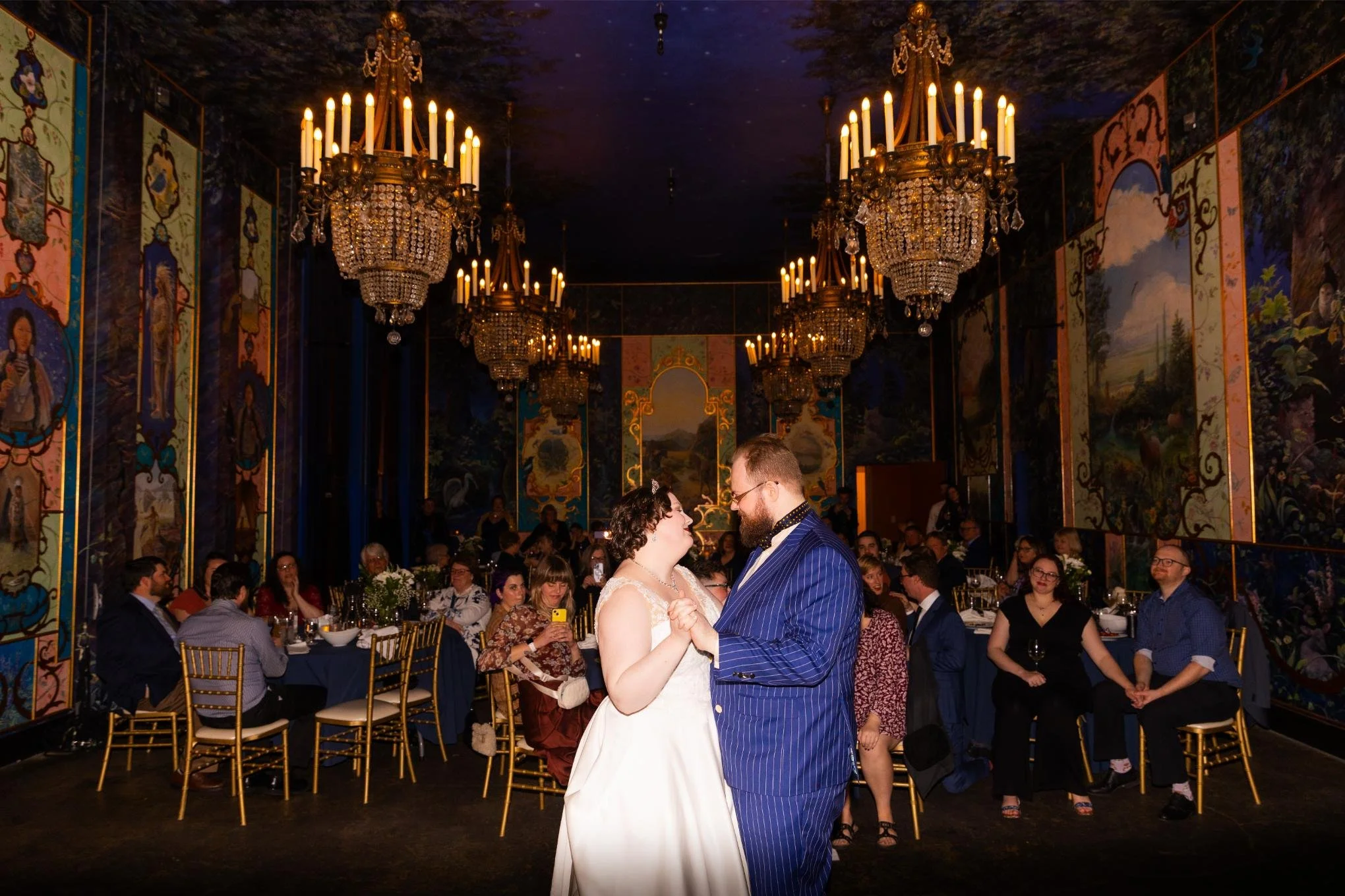 A bride and groom dance closely at their wedding reception at The Ruins in Seattle in an elegant venue with large chandeliers and colorful murals on the walls. Guests sit at decorated tables watching the couple.