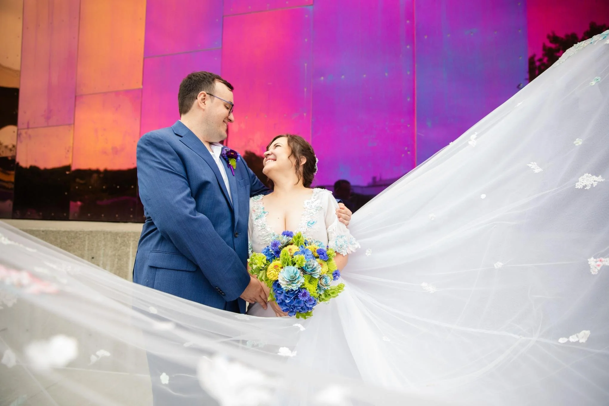 A bride and groom sharing a moment of happiness with a backdrop of colorful sunset reflected on a modern building. The bride is holding a bouquet of vibrant flowers, and they are holding hands with the bride's wedding dress flowing around them.
