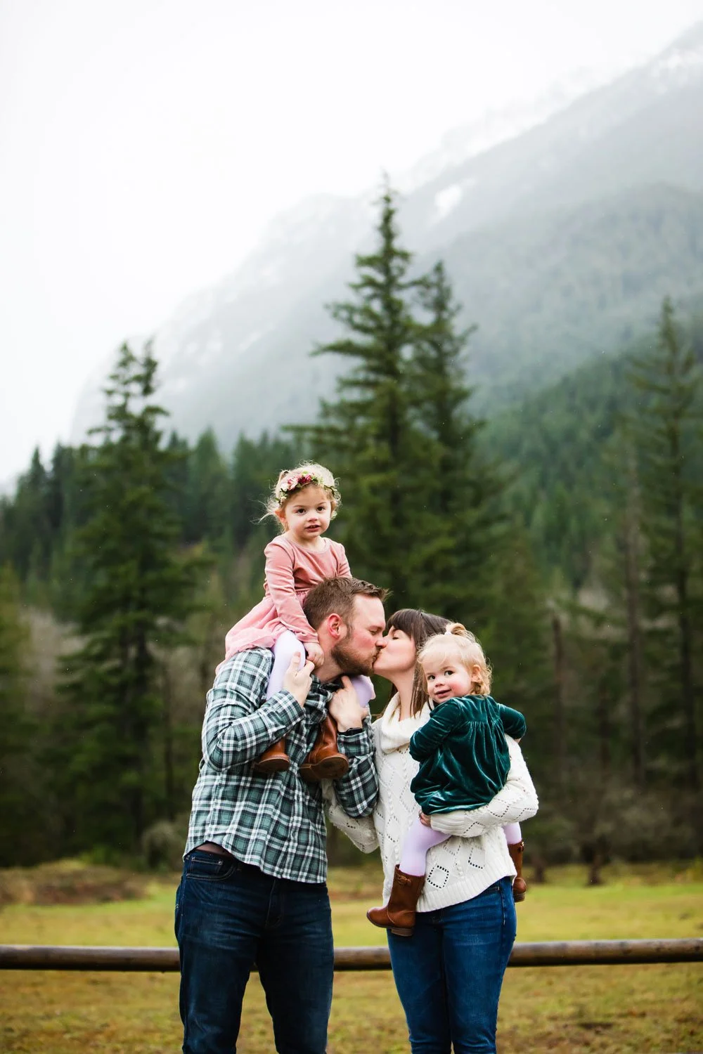 A family of four outdoors in front of a forest and mountain backdrop, sharing a kiss with two young girls on their shoulders.
