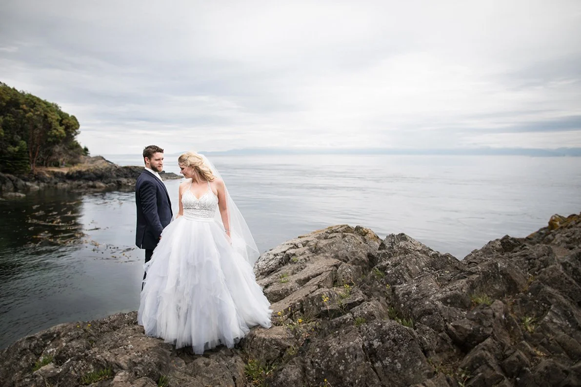 A bride and groom standing on rocks by the water at Lime Kiln on San Juan Island with a cloudy sky in the background.