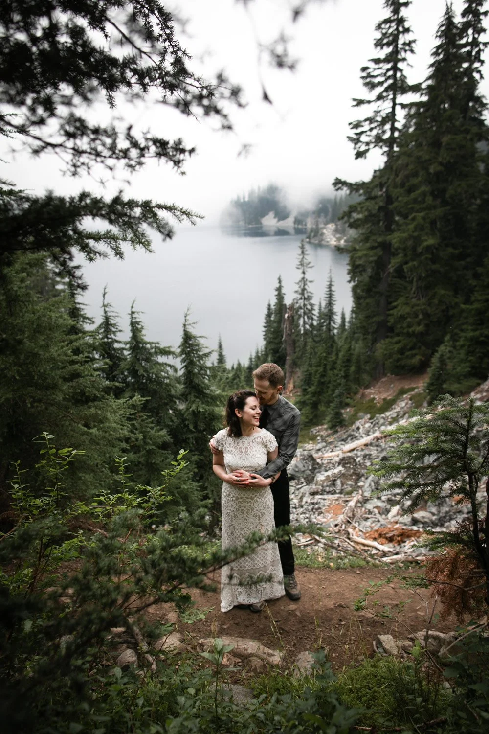 A couple stands close together in a forested area near a lake, with tall pine trees and foggy mountains in the background while smiling and sharing a moment.