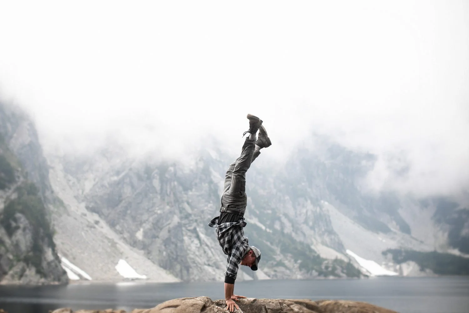 Person performing a handstand on a rock near a lake with mountainous landscape and mist in the background.