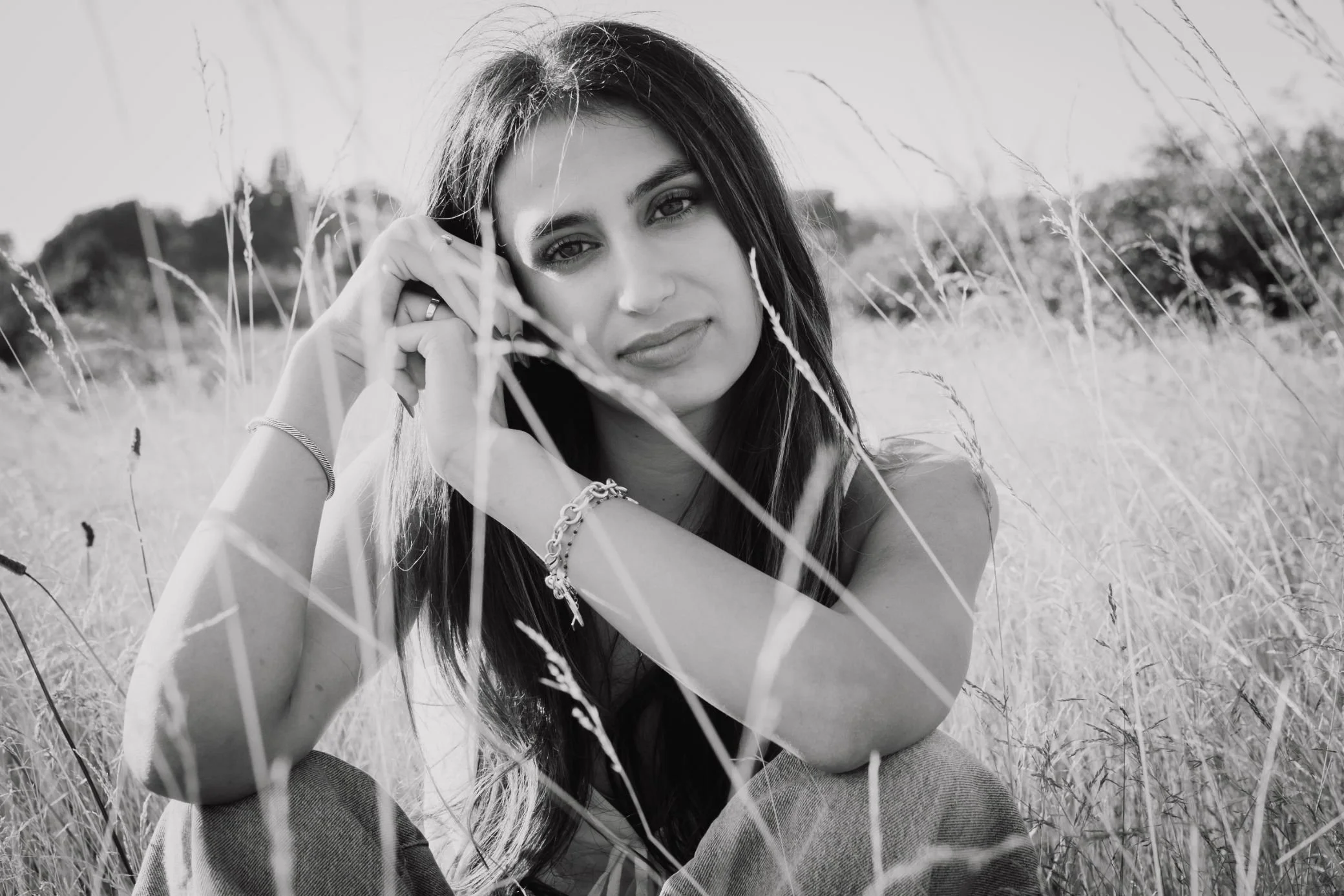 A young woman with long hair and jewelry sitting in a field of tall grass, looking at the camera in black and white.