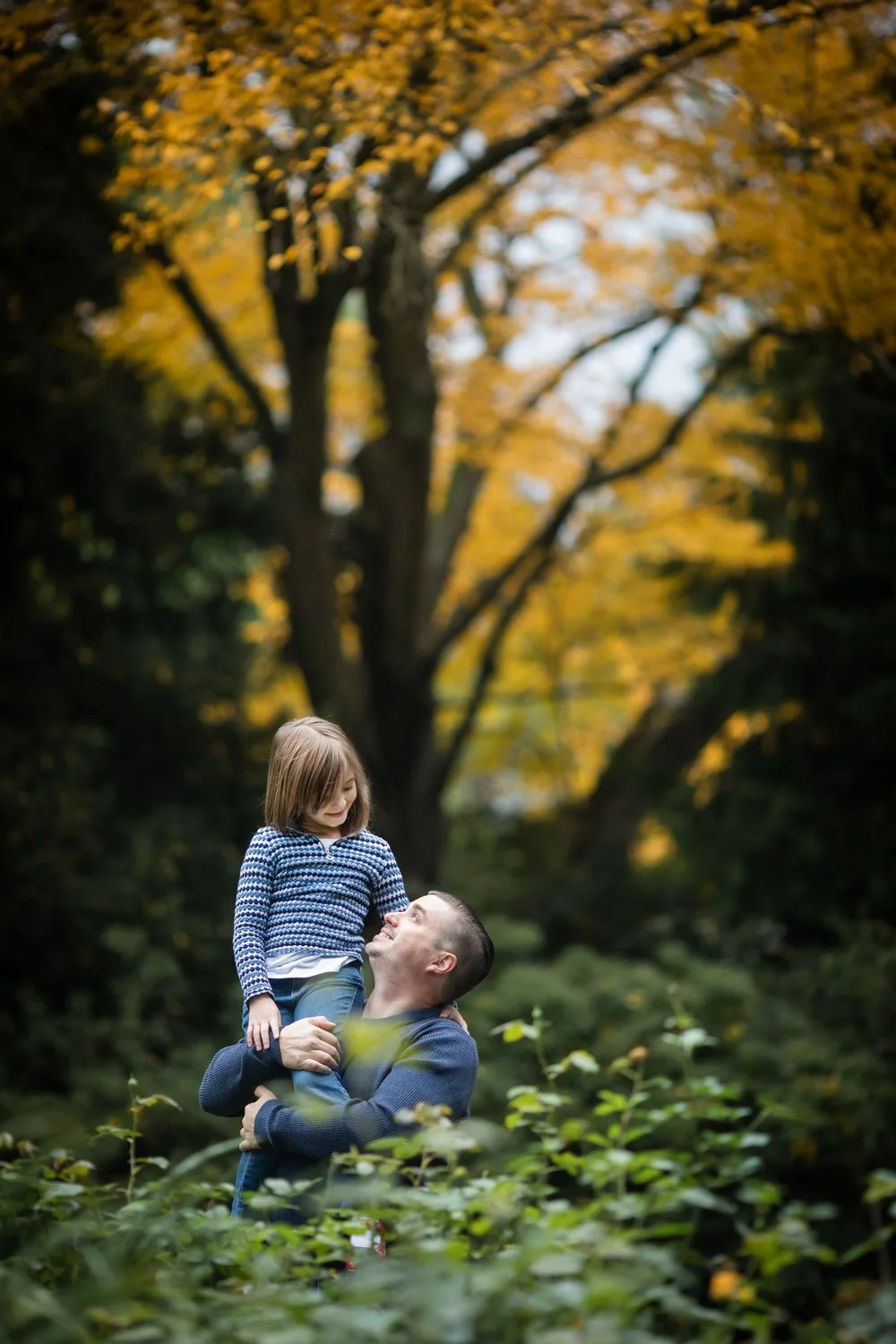 A man holding a young girl in his arms outdoors with autumn trees in the background.