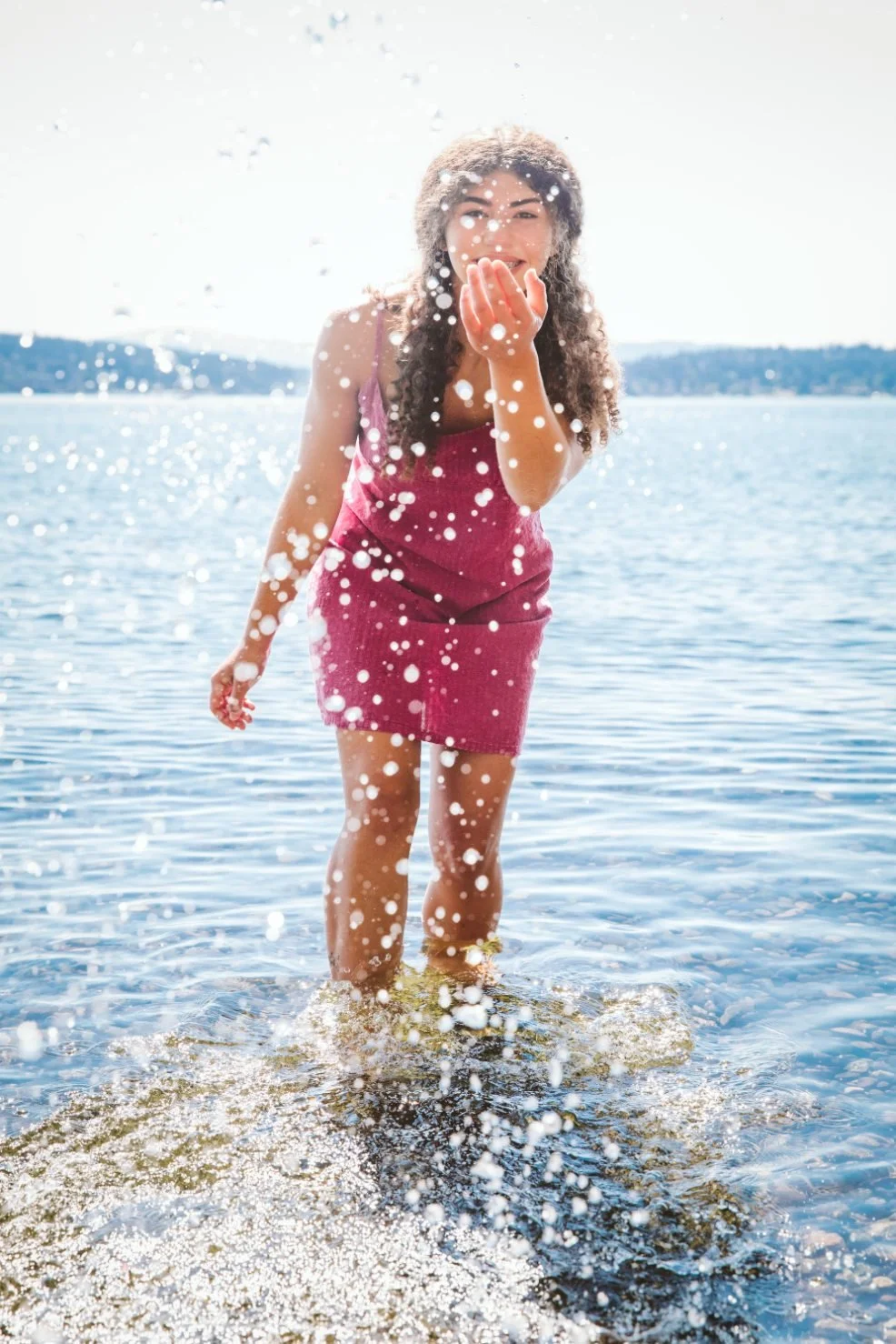 Woman in pink dress playing and celebrating in water at the beach on a sunny day, splashing water