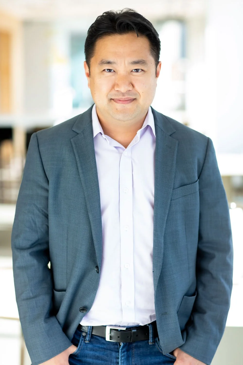 A man wearing a dark gray suit jacket and white shirt standing indoors with arms relaxed, smiling, with a blurred modern office background.