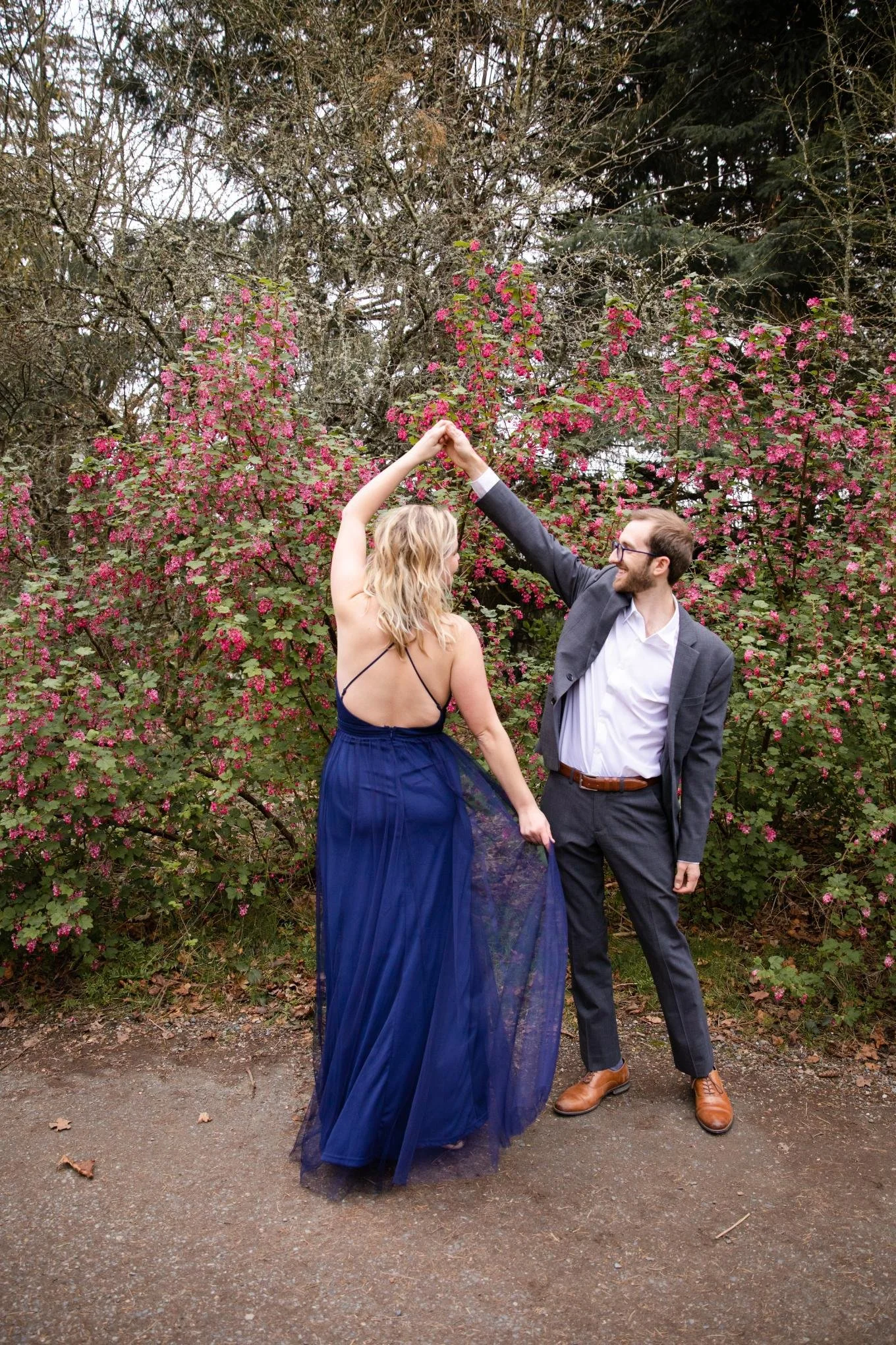 A man and woman dancing outdoors in front of flowering bushes. The woman wears a blue dress, and the man is dressed in a suit.