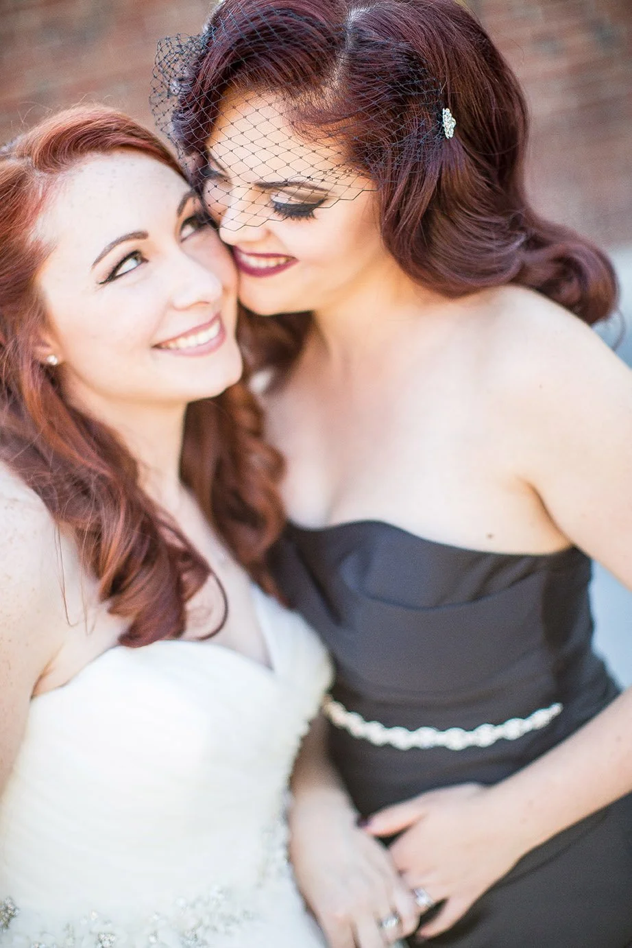 Two women with red hair smiling close together, one in a white dress and the other in a black dress with a veil and hair clip, embracing each other affectionately.