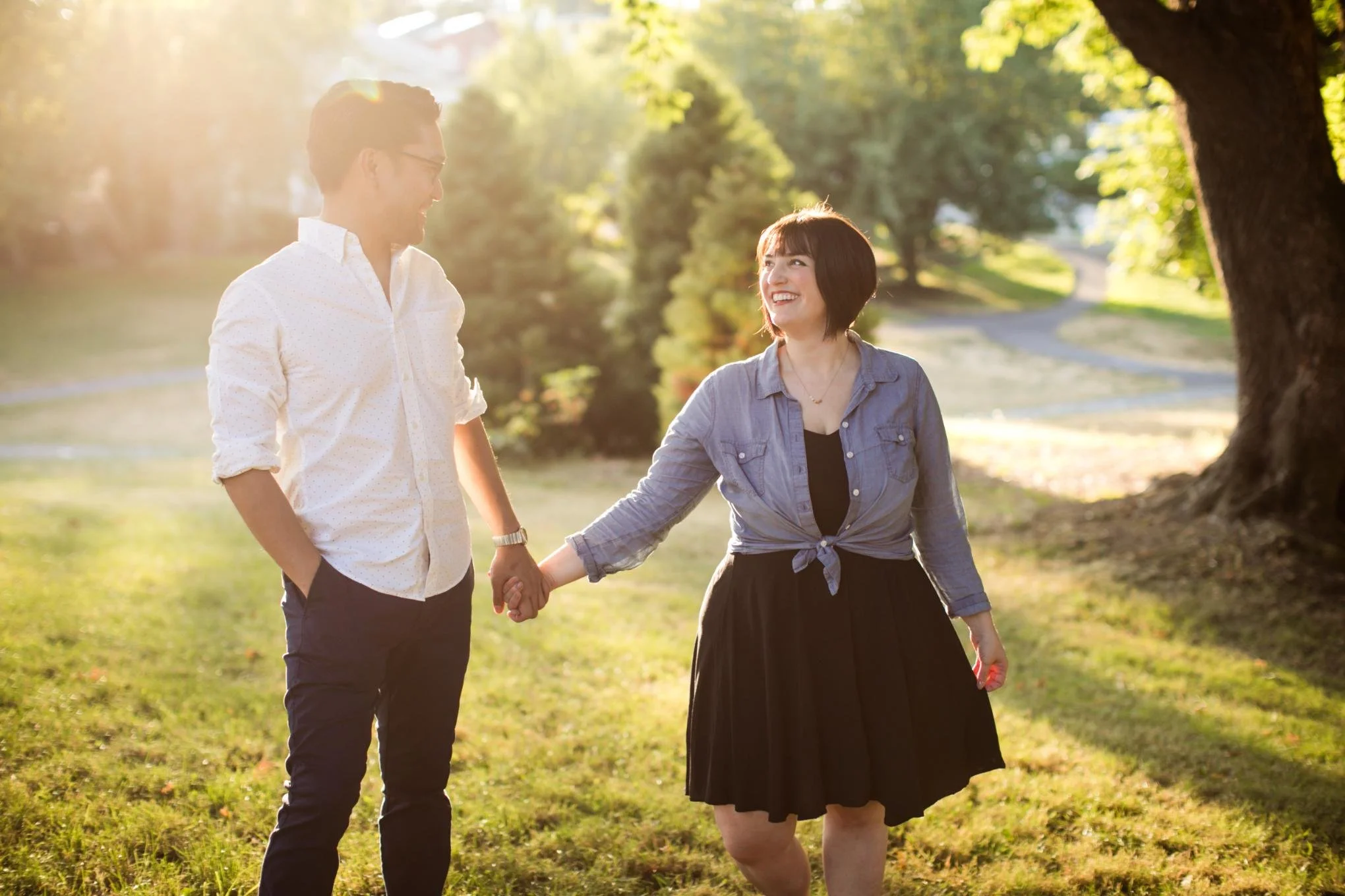 A couple holding hands and smiling in a park with trees in the background, during the golden hour.