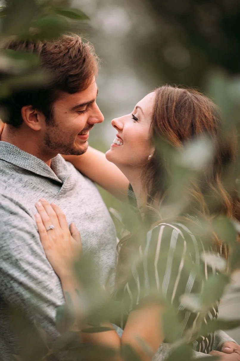 A young couple smiling and gazing into each other's eyes outdoors, surrounded by blurred greenery.