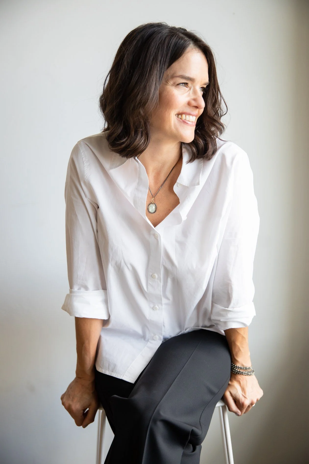 A woman with dark brown, wavy hair, smiling and looking to her right, wearing a white button-up shirt with rolled-up sleeves, black pants, and a pendant necklace, sitting on a stool against a plain background.