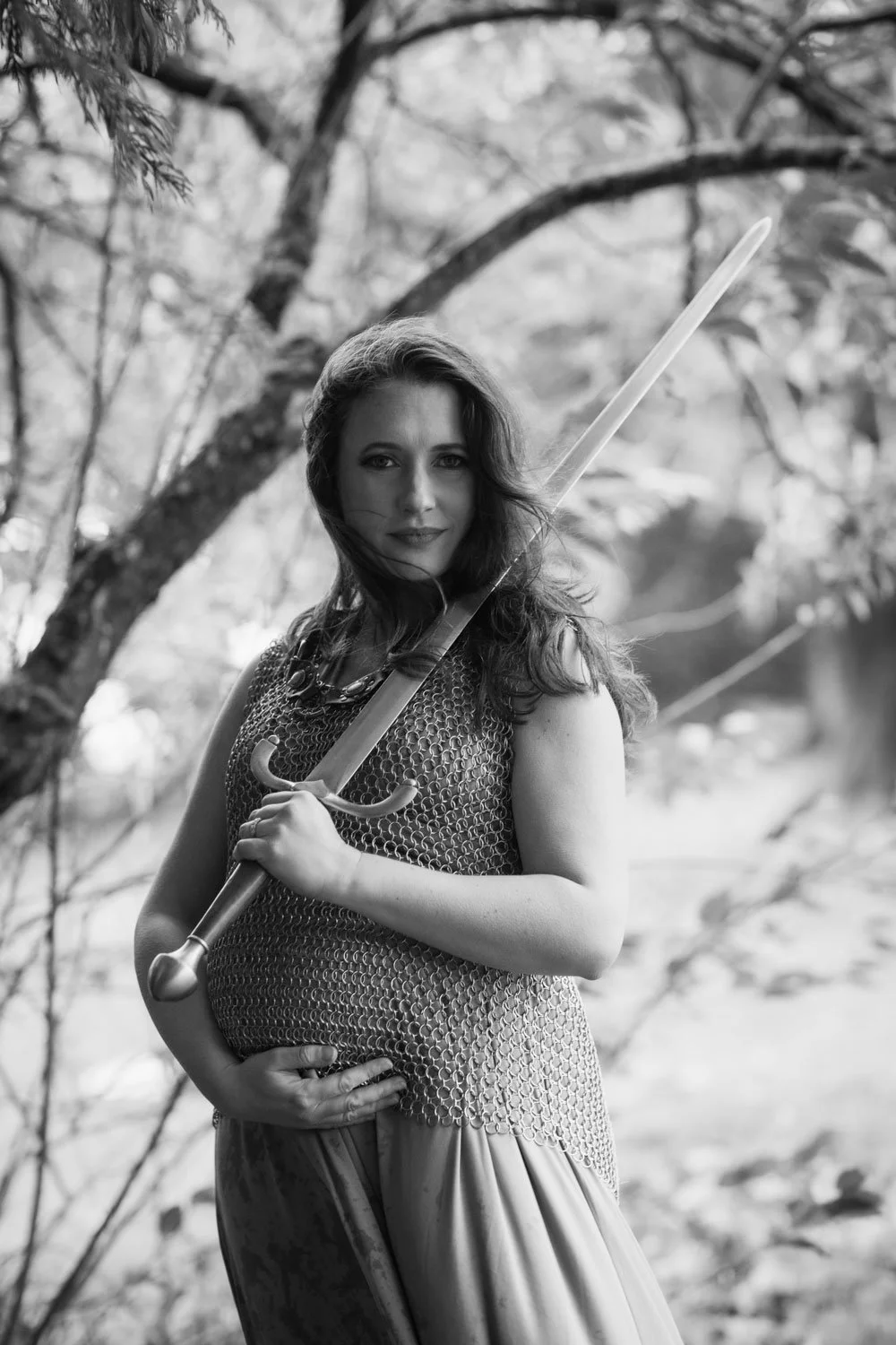 A pregnant woman in medieval armor holding a sword outdoors with trees in the background.
