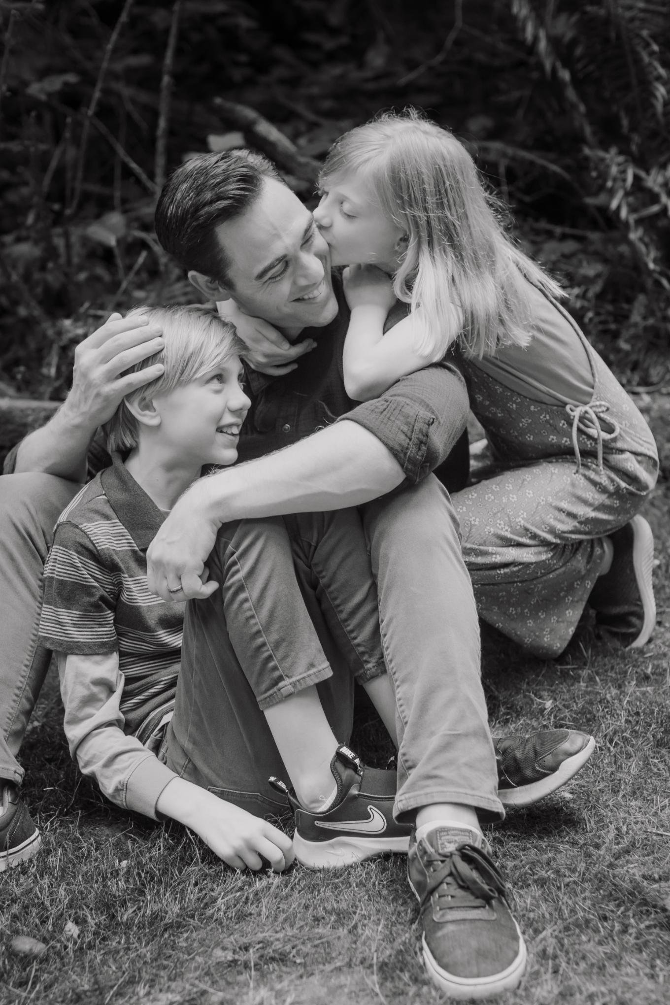 A family outdoors, including a man, a girl, and a boy, sharing a joyful moment. The girl is giving the man a kiss on the cheek, and the boy is looking up at the man with a smile.