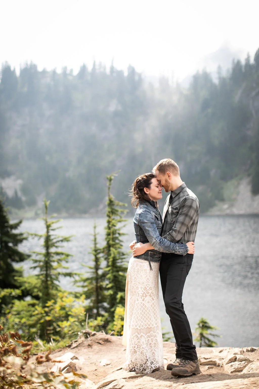 A couple embracing near a lake with a forested mountain background.