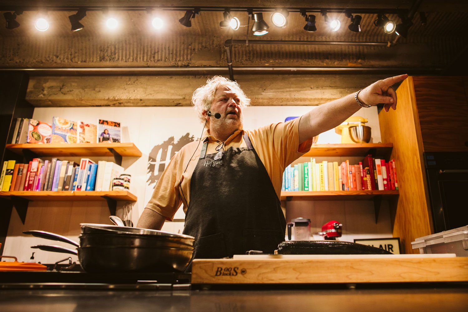 A man Tom Douglas with gray hair and a beard, wearing a beige shirt and black apron, is standing in a kitchen and pointing to the right. He is wearing a headset microphone, and there are cookbooks and kitchen appliances behind him.