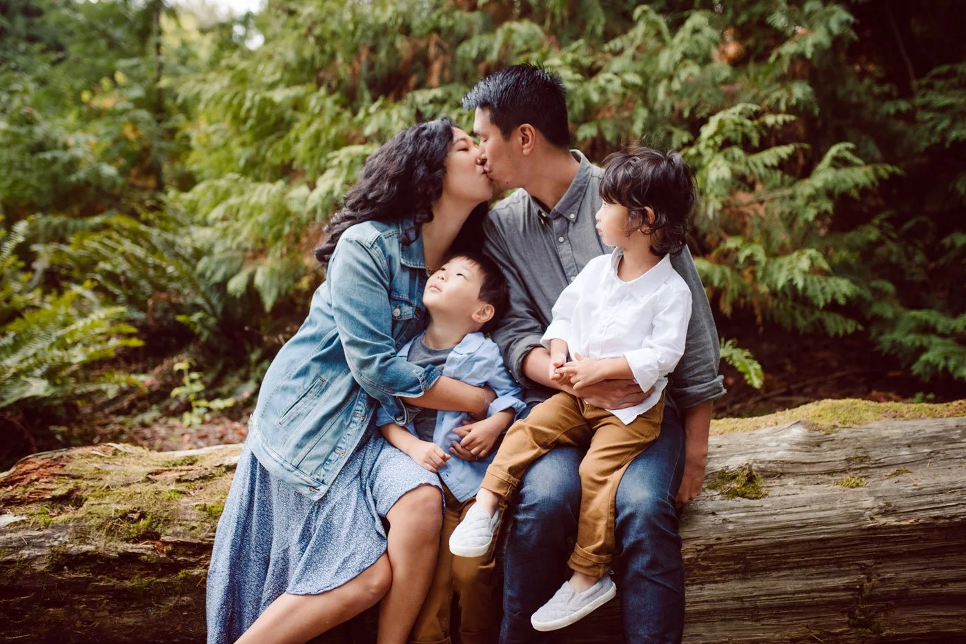 Family of four sitting on a log in a forest, sharing a kiss while the children watch, surrounded by green foliage.