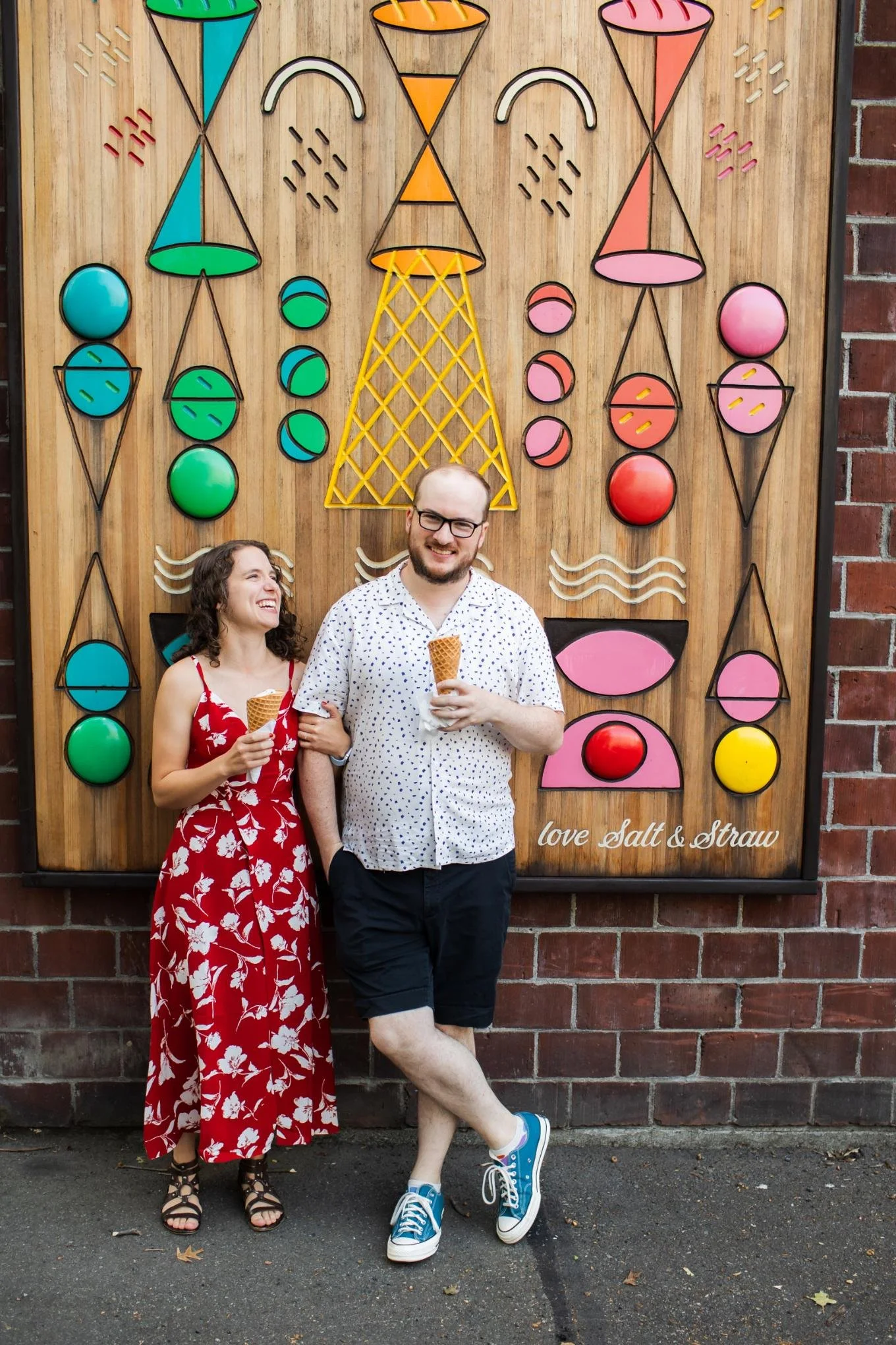 A smiling woman in a red floral dress and a smiling man in a white polka dot shirt and black shorts are standing in front of a colorful wooden art installation, holding ice cream cones.
