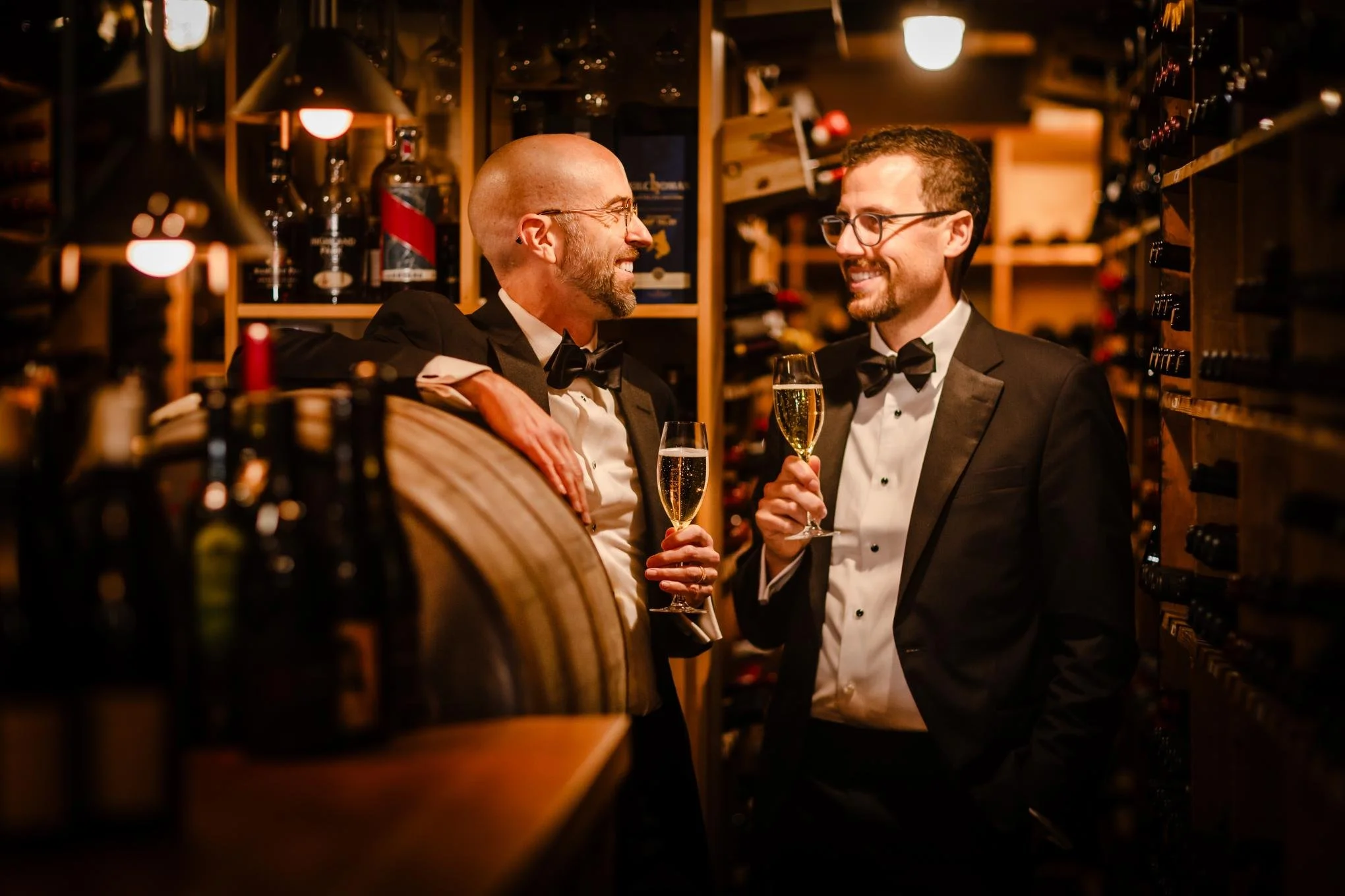 Two men in tuxedos sharing a toast with champagne in a wine cellar, smiling and looking at each other.
