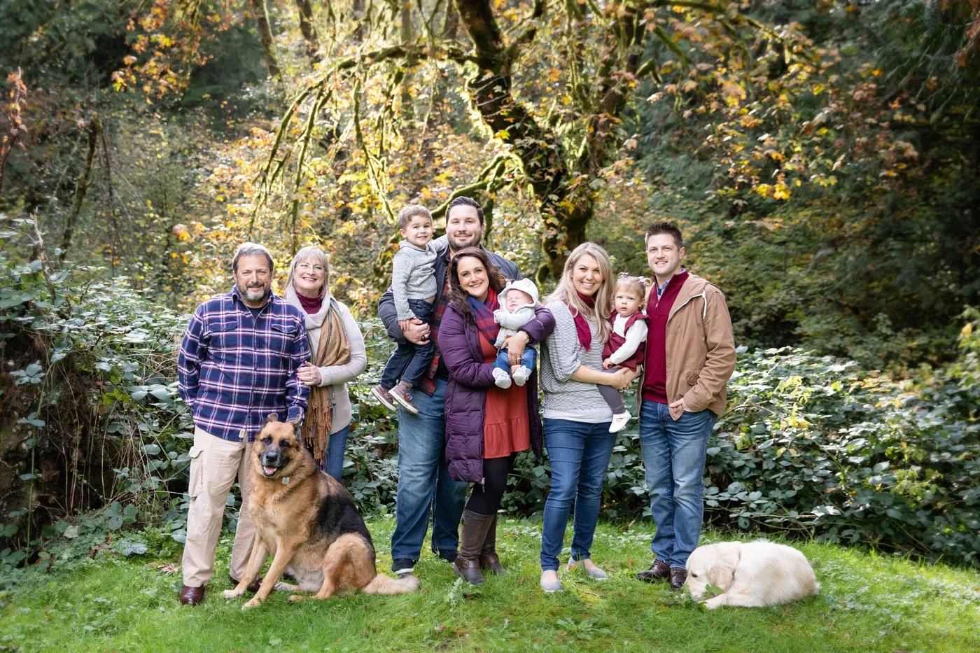 Family with three adults, two children, and two dogs standing in a wooded outdoor area during fall.