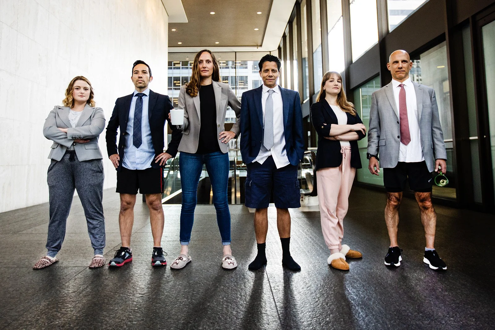 Six professionally dressed people standing in a line inside a modern office building, wearing pajama bottoms and looking confidently at the camera.